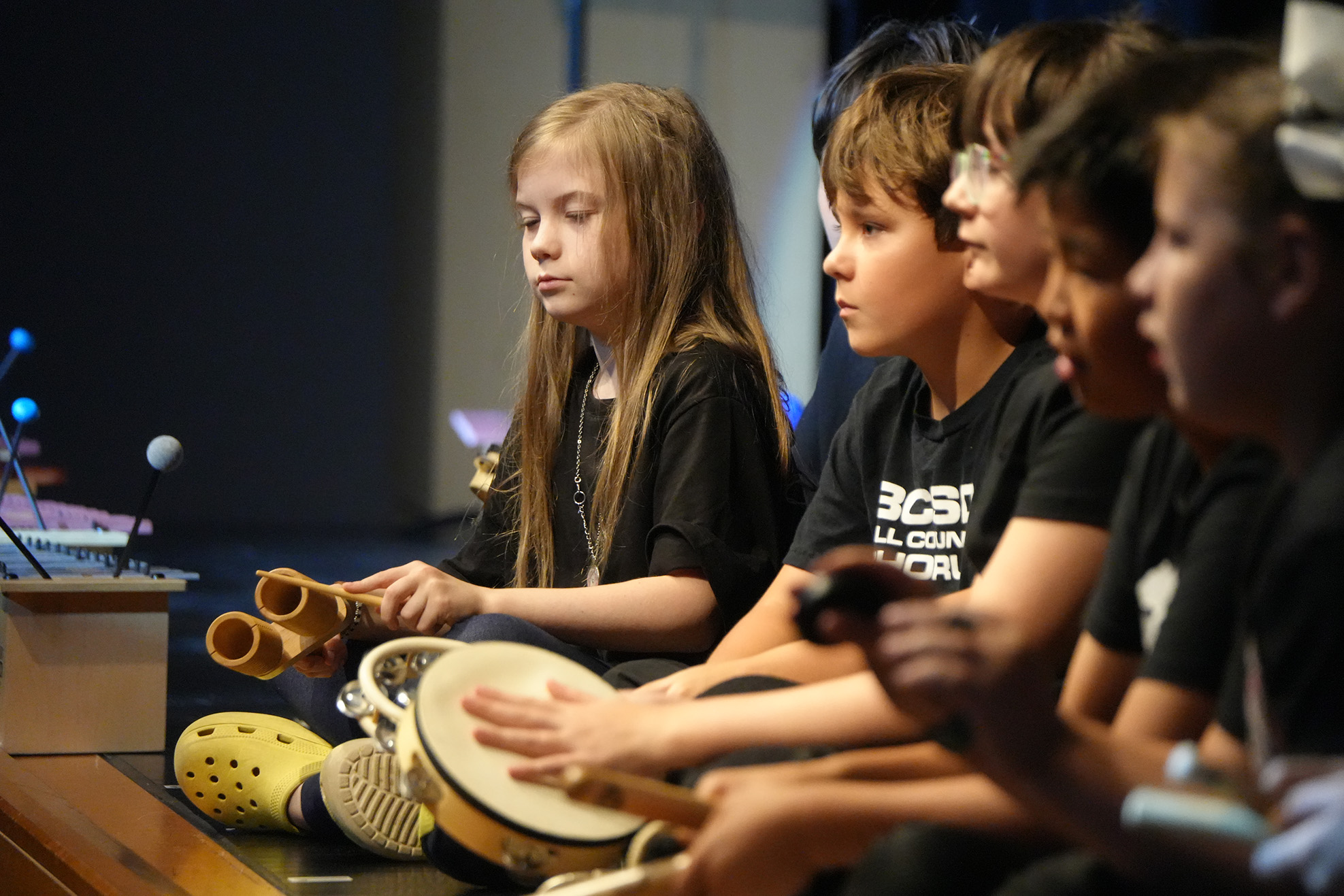 Four kids wearing black T-shirts sit in a row, one playing a drum, another a marimba.