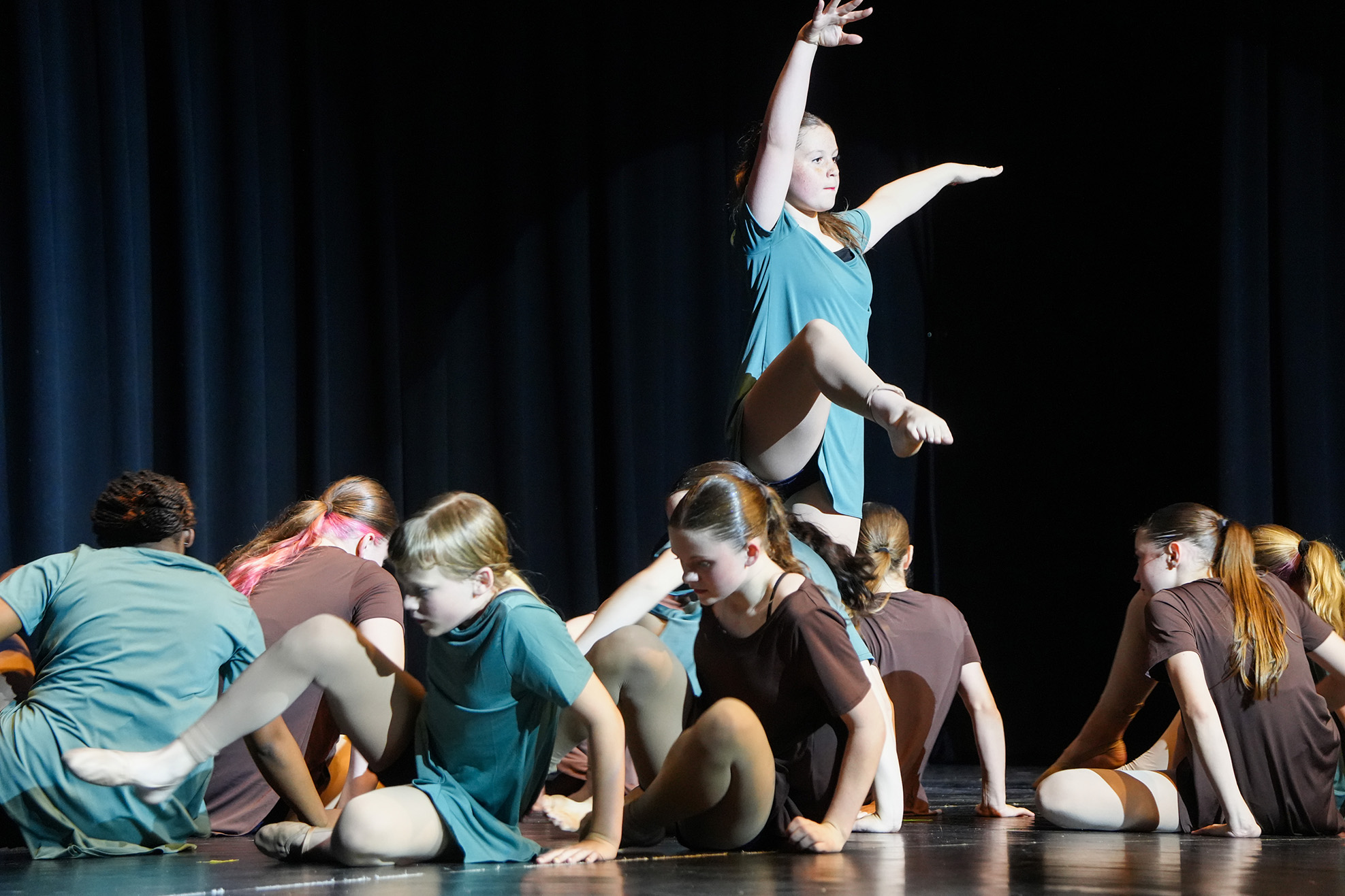 A group of dancers on stage, one woman jumping, others seated in various poses, against a dark background.