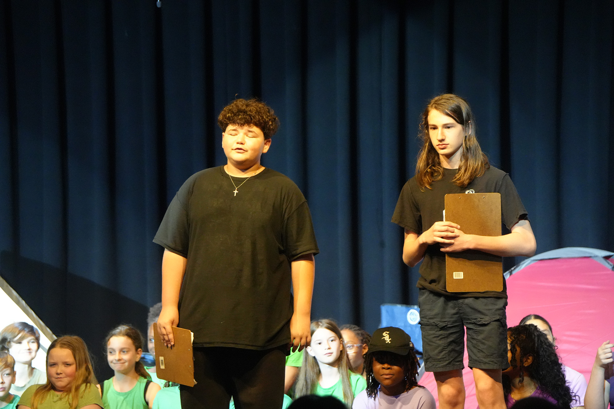 Two young people on stage holding clipboards, with a blue curtain in the background.