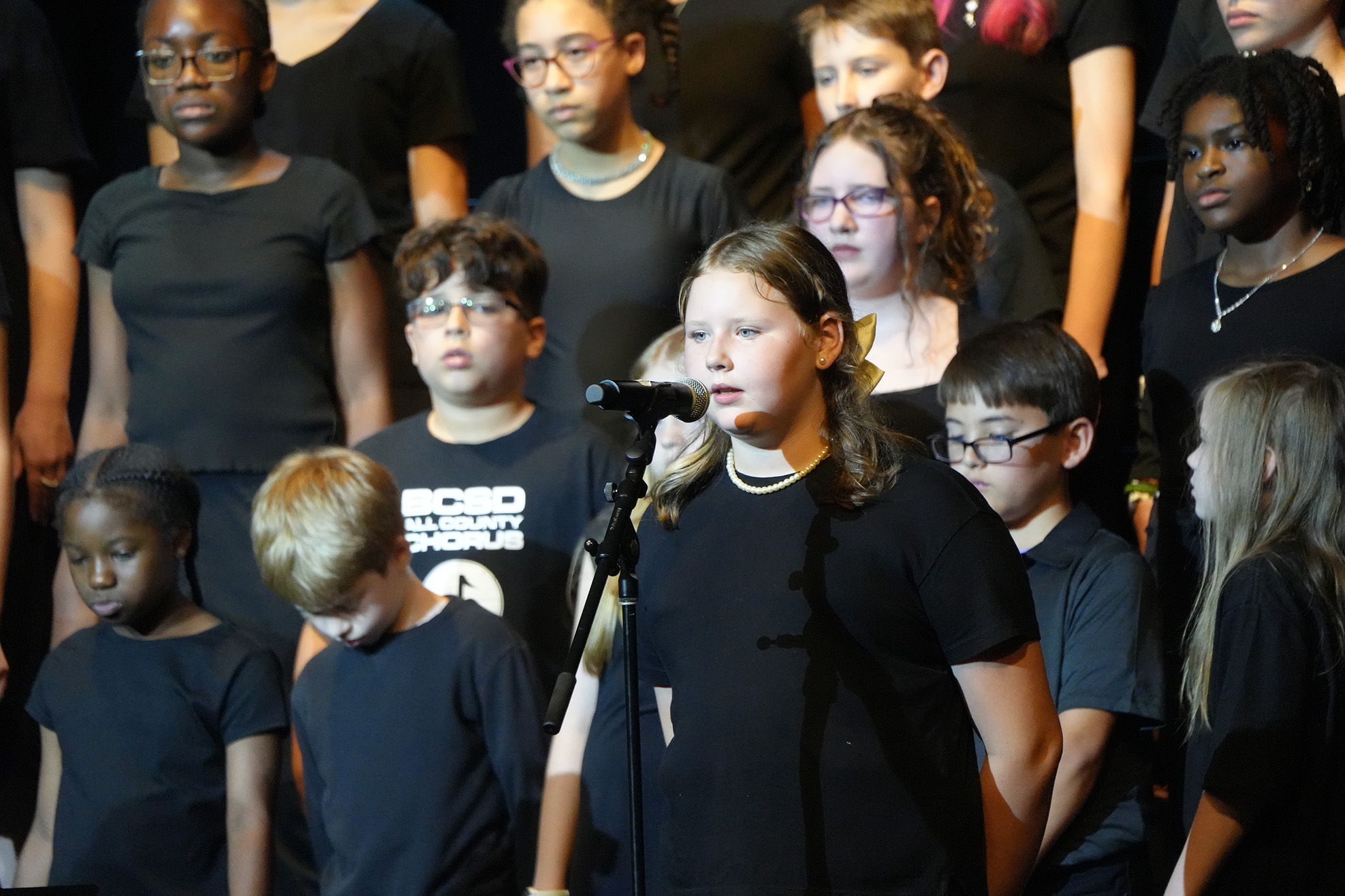 A choir of children in black t-shirts performing on stage, with one child singing into a microphone.