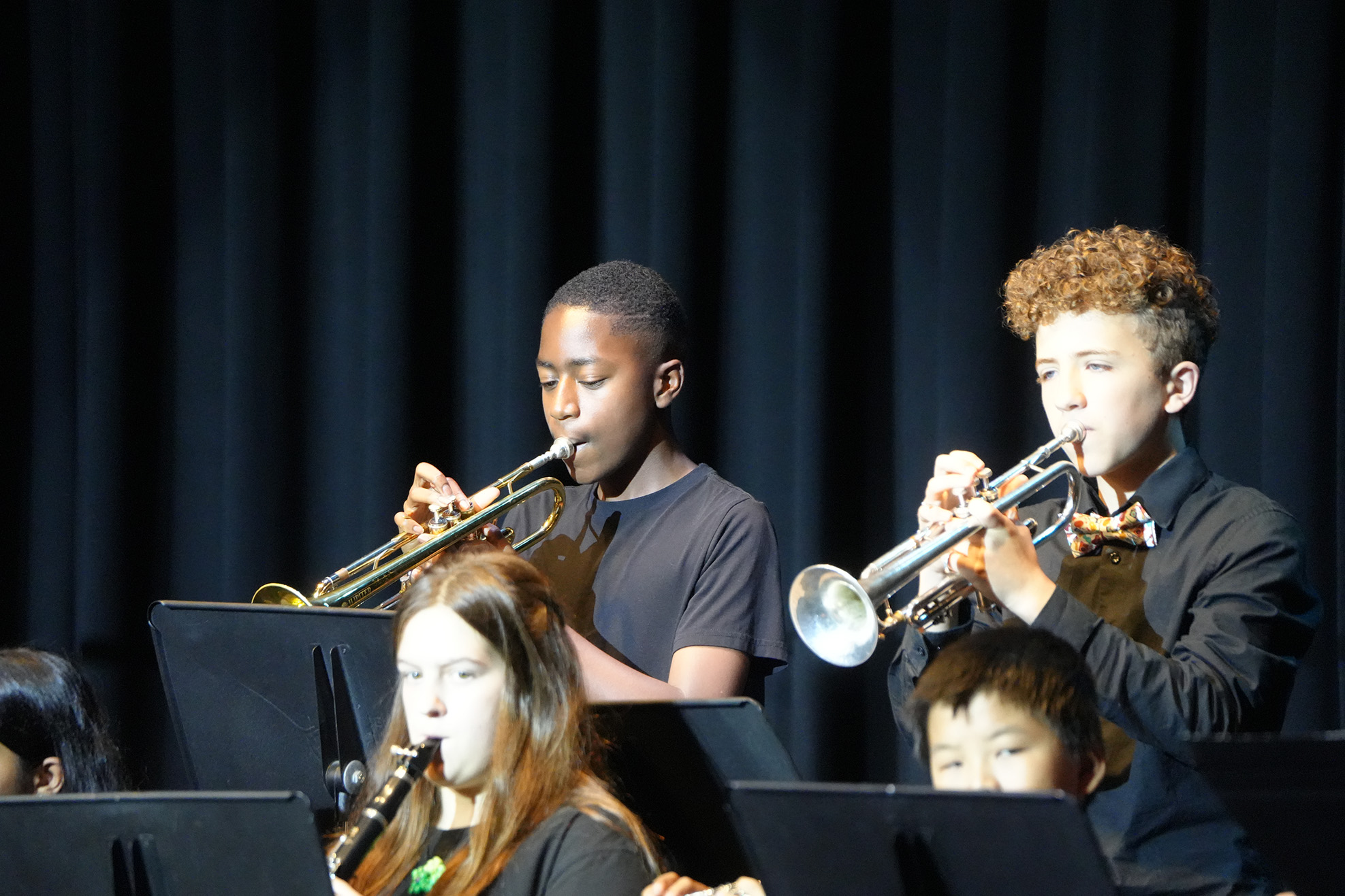 Children play musical instruments in a music room. Two boys play trumpets; one wears a bow tie. A girl plays a clarinet.