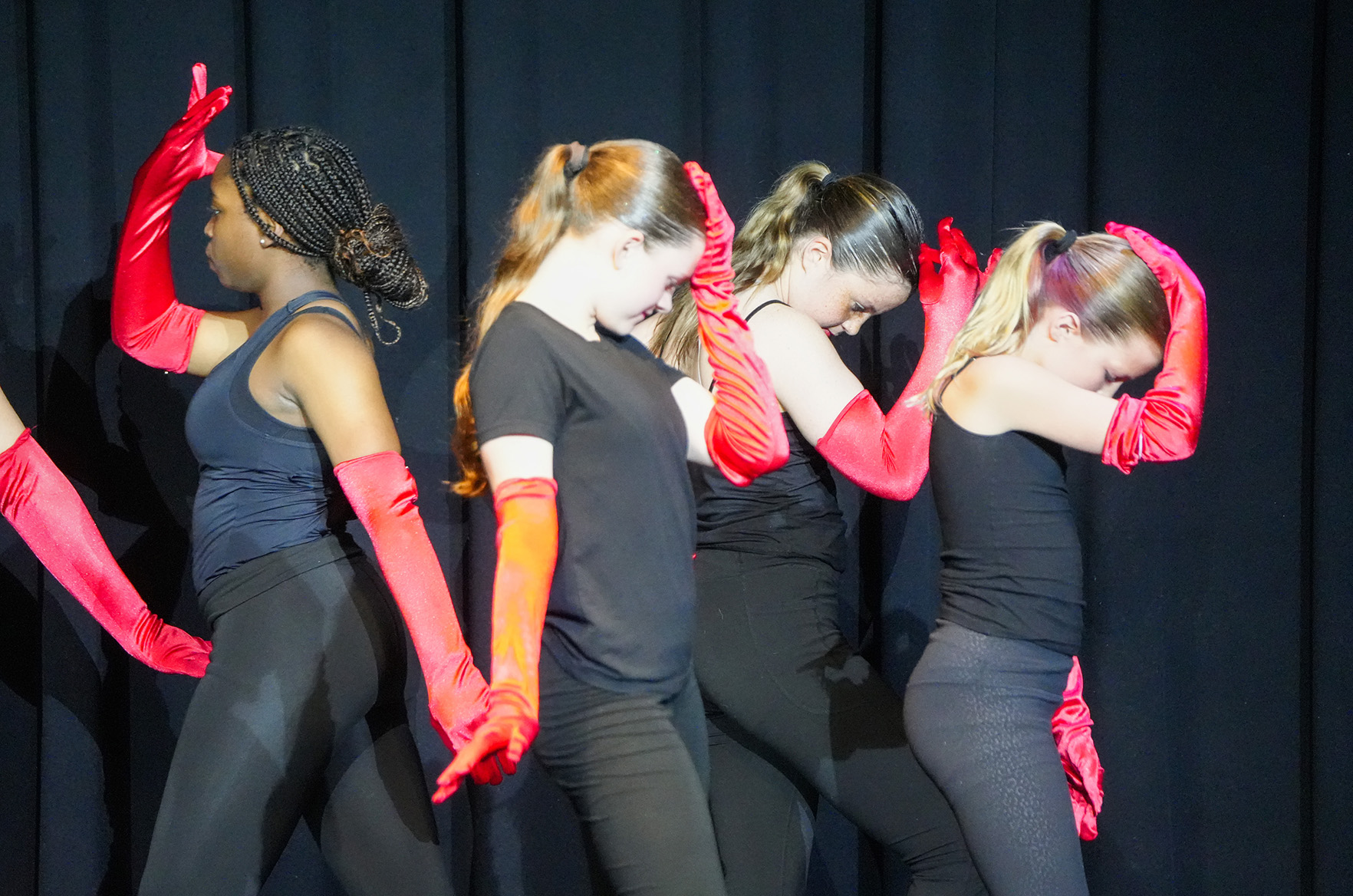 Five women on stage wearing black tops and leggings, red gloves, and wigs, performing a dance routine.