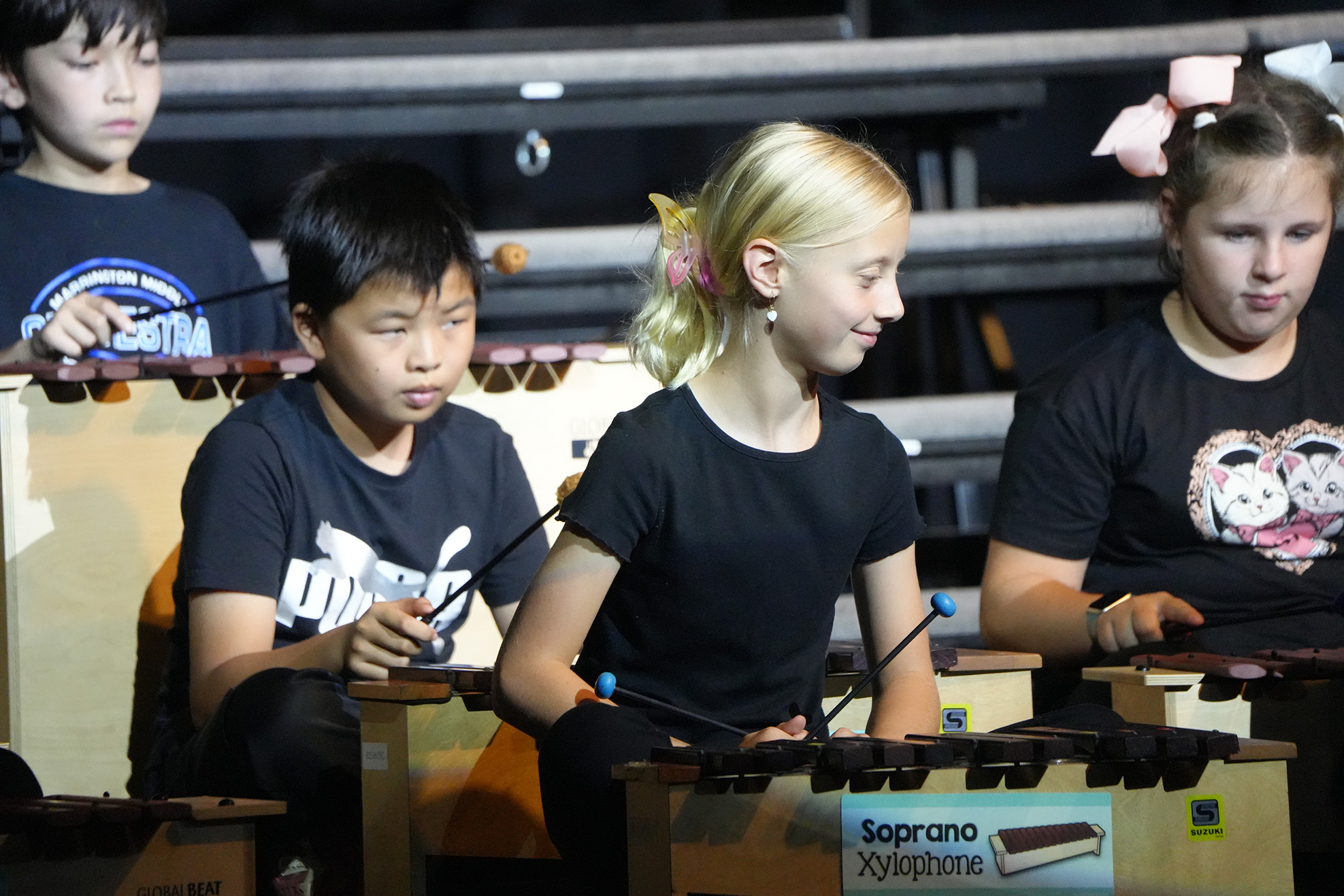 Four children in black t-shirts play xylophones in a stadium. Each child holds mallets.