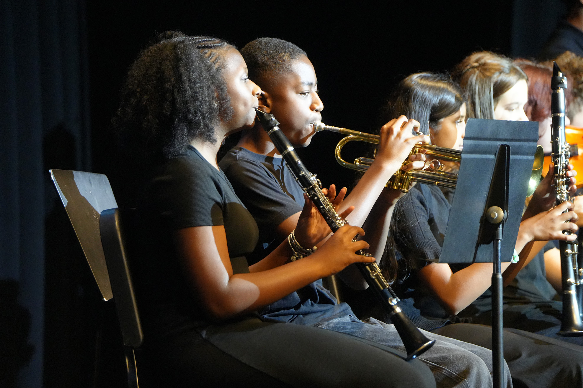 A group of young musicians sit on stage. They play trumpet, clarinet, and oboe. Behind them, a dark backdrop.