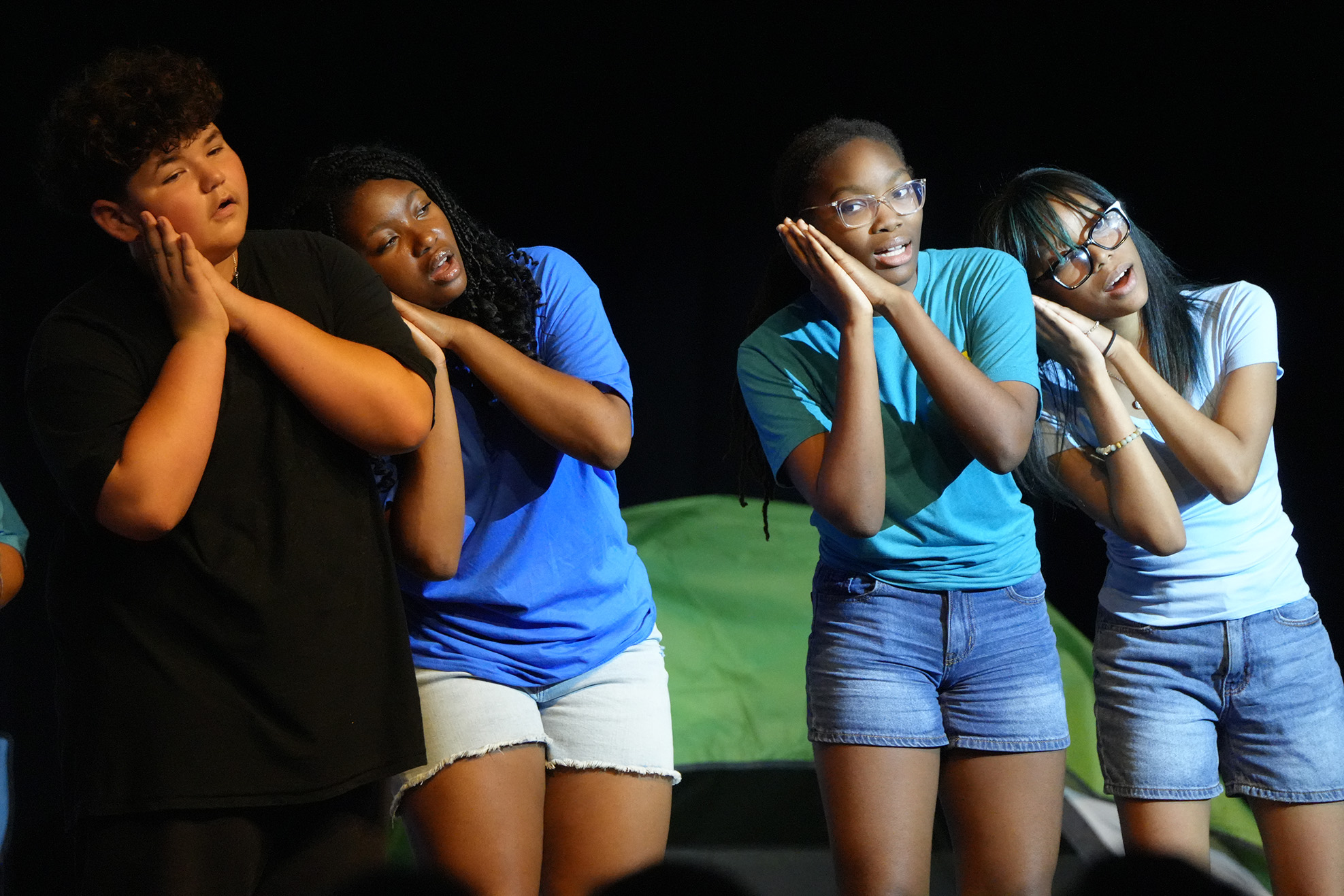 Four individuals in casual attire stand on a stage with hands over their mouths. The background features a green tent.