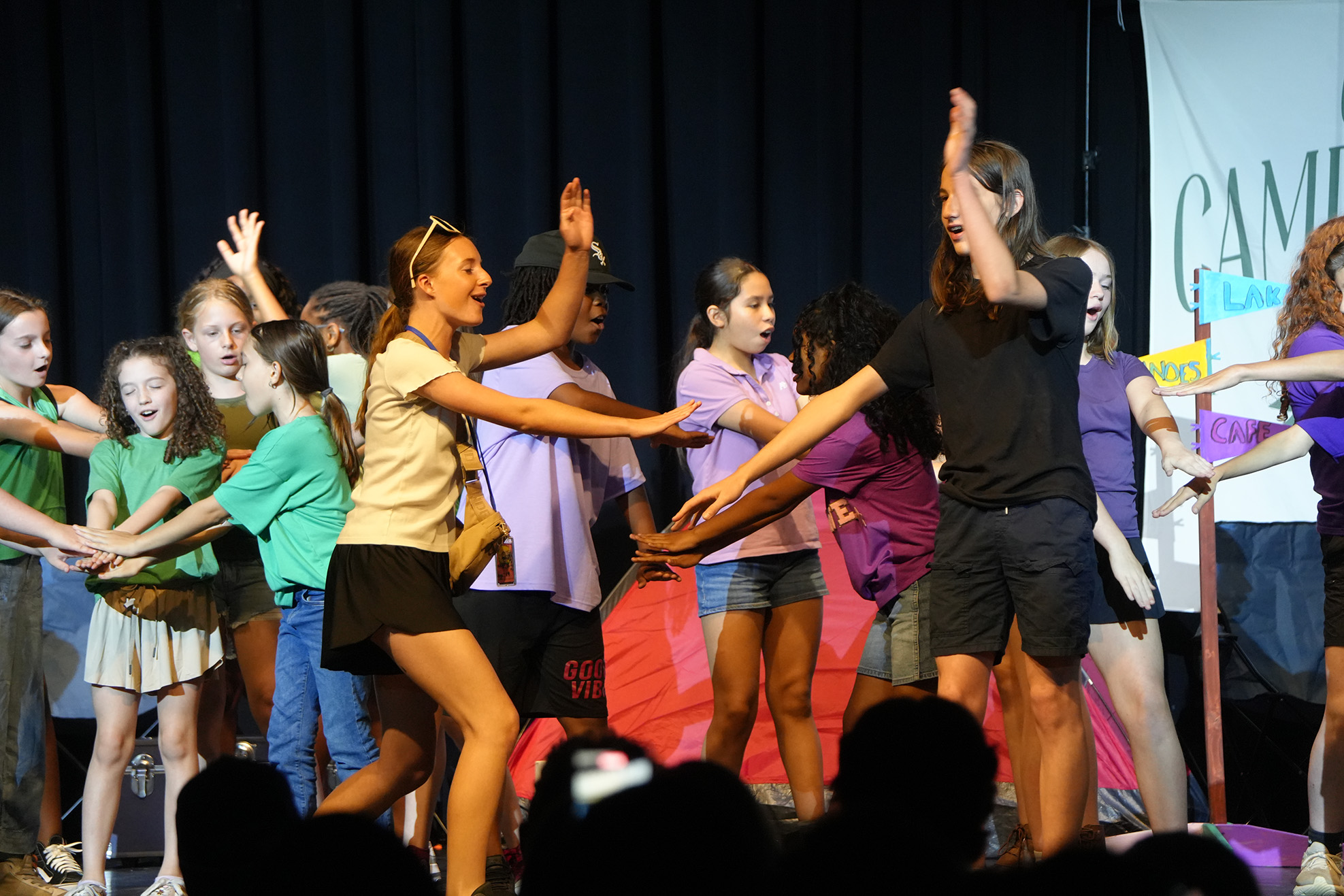 A group of children performing on stage, some with hands raised, wearing varied summer clothing.