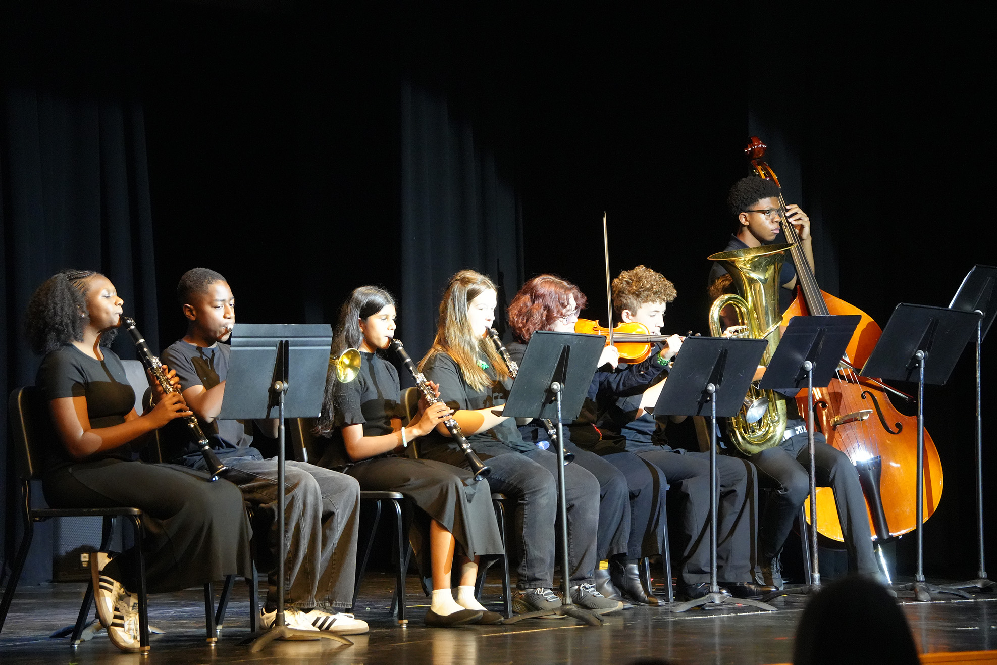 A group of students perform on instruments on stage, including a violin, a cello, and clarinets.