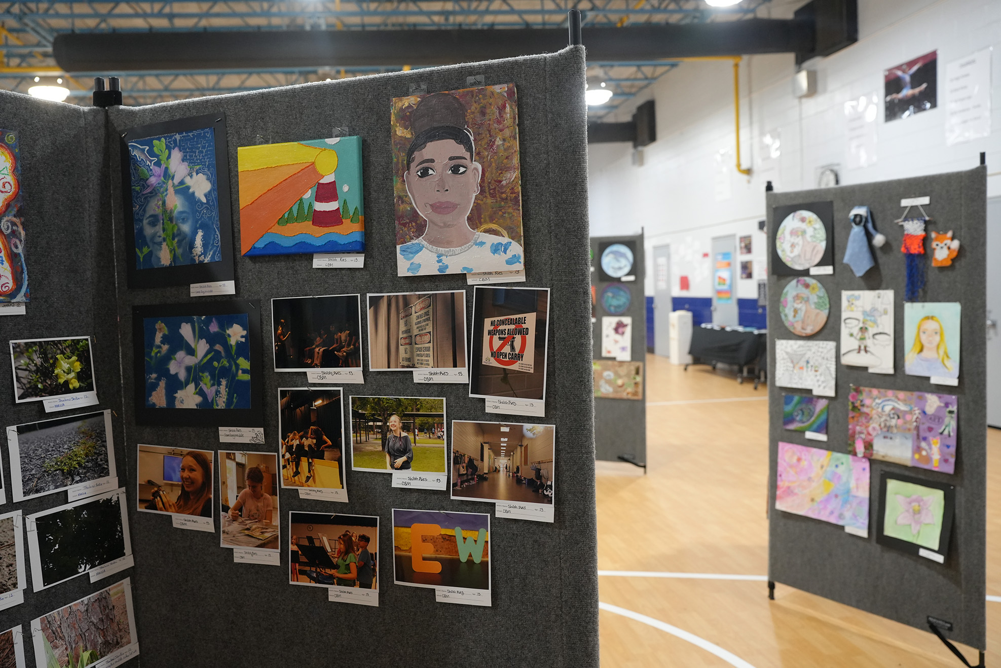 Display of various artwork on boards in a gym with wooden floor, showcasing diverse art styles and subjects.