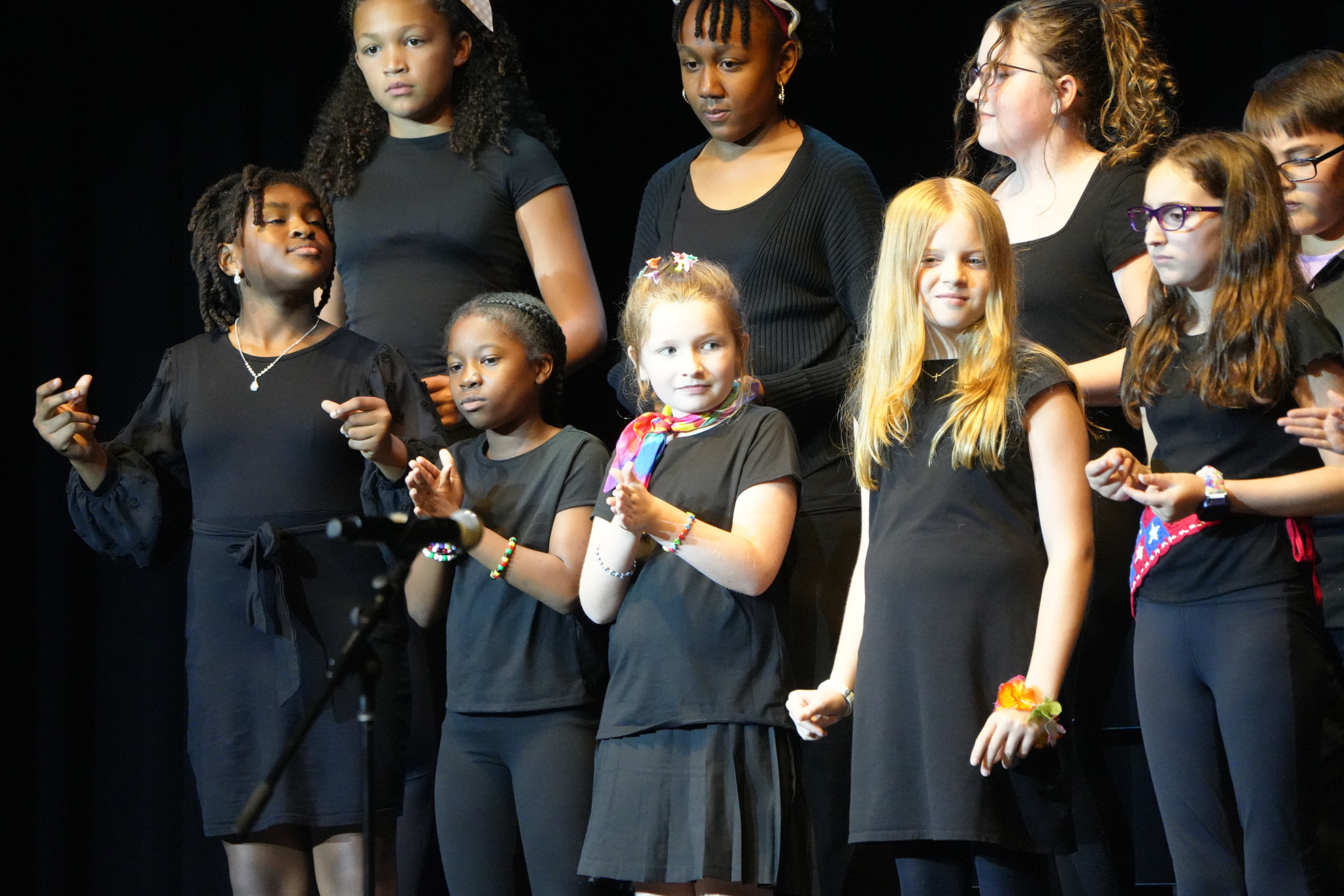 A group of young people in black clothes on a stage, some clapping, with a black backdrop.