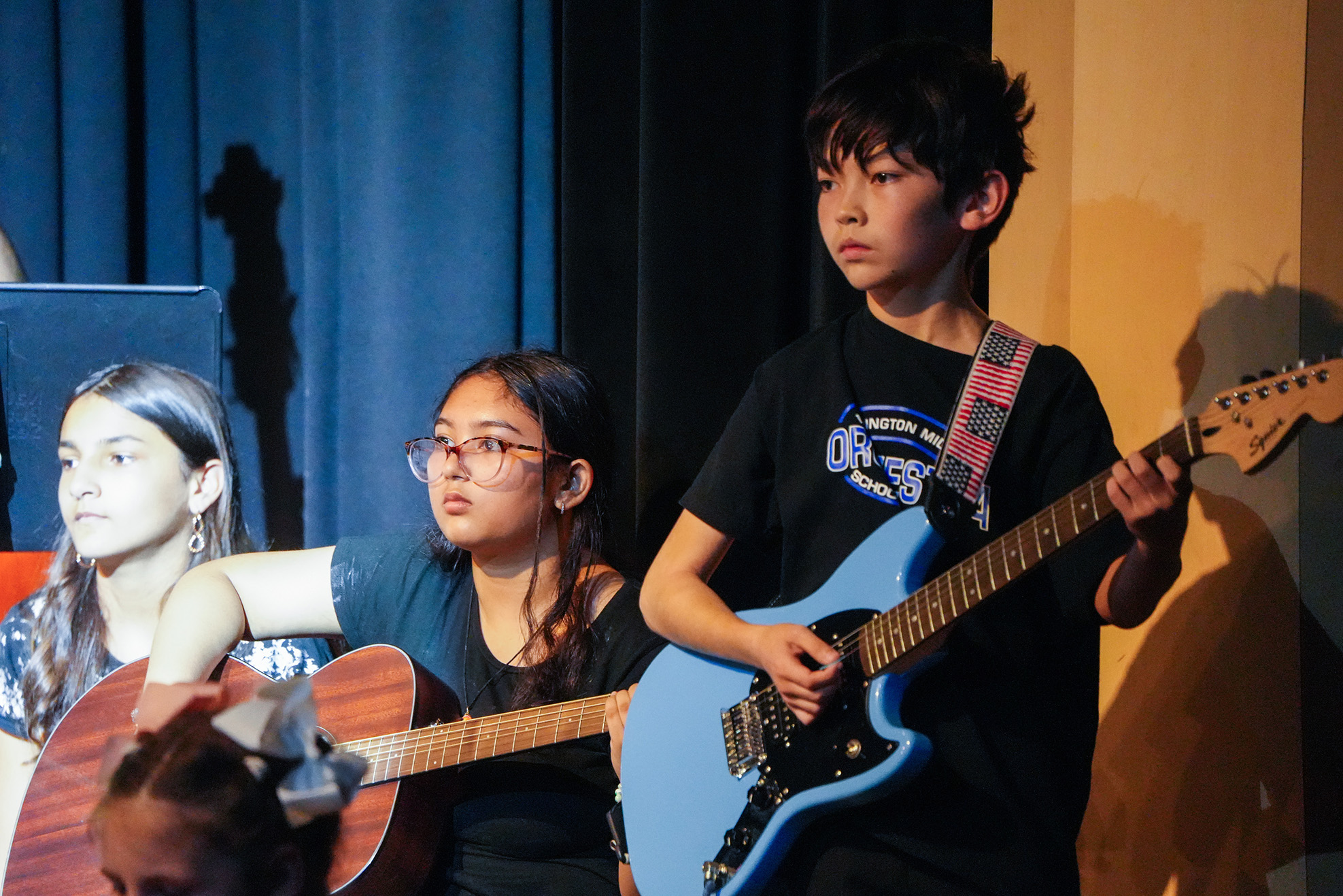 Three young musicians stand on stage, each holding a guitar. One plays a blue electric guitar, another a brown acoustic, and the third waits with a guitar.
