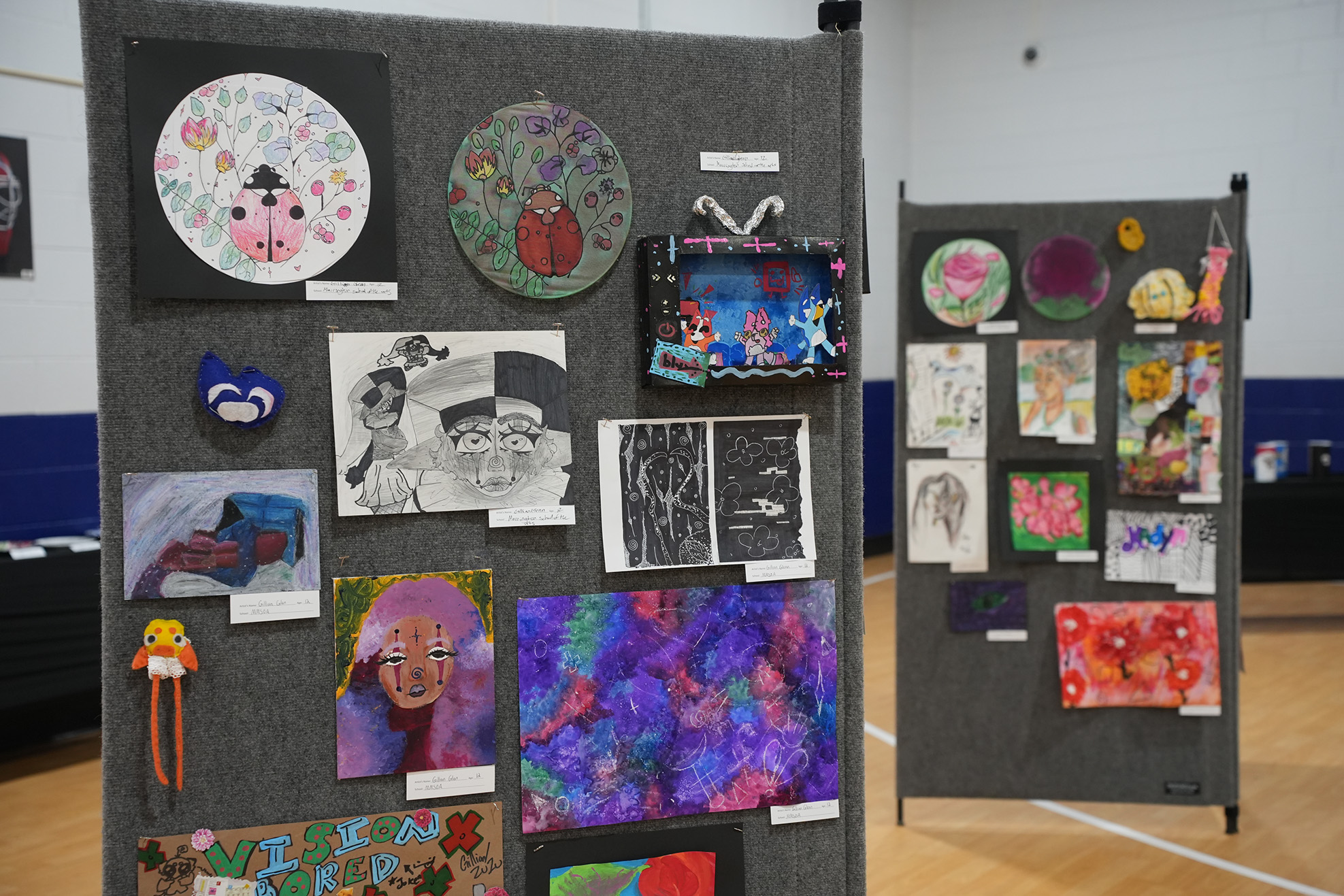 Multiple art pieces on a display board, including drawings, paintings, and a red box. Background is a gym.