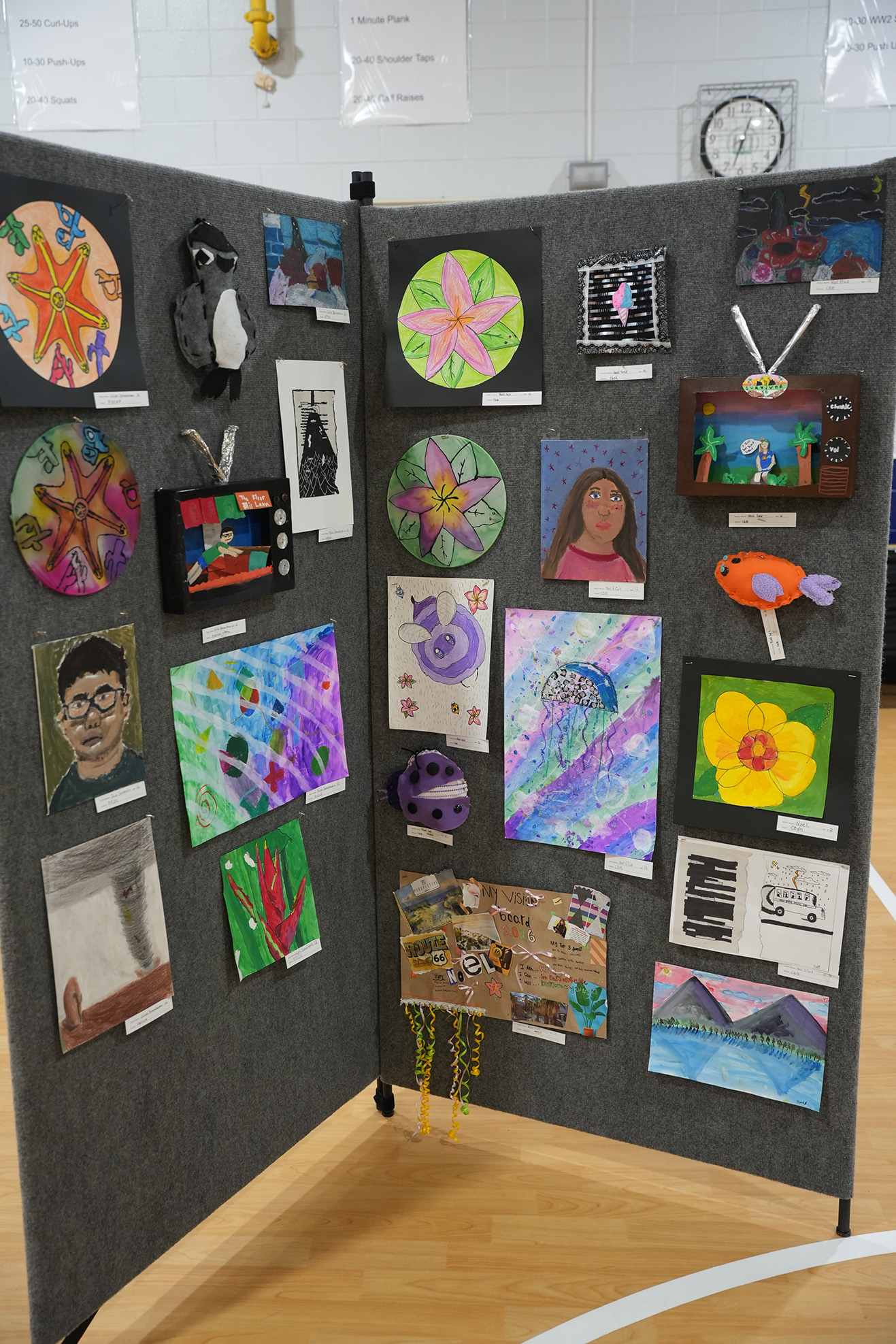 Display of student artwork on a gray wall with wooden floor. Pieces include portraits, flowers, and collages.