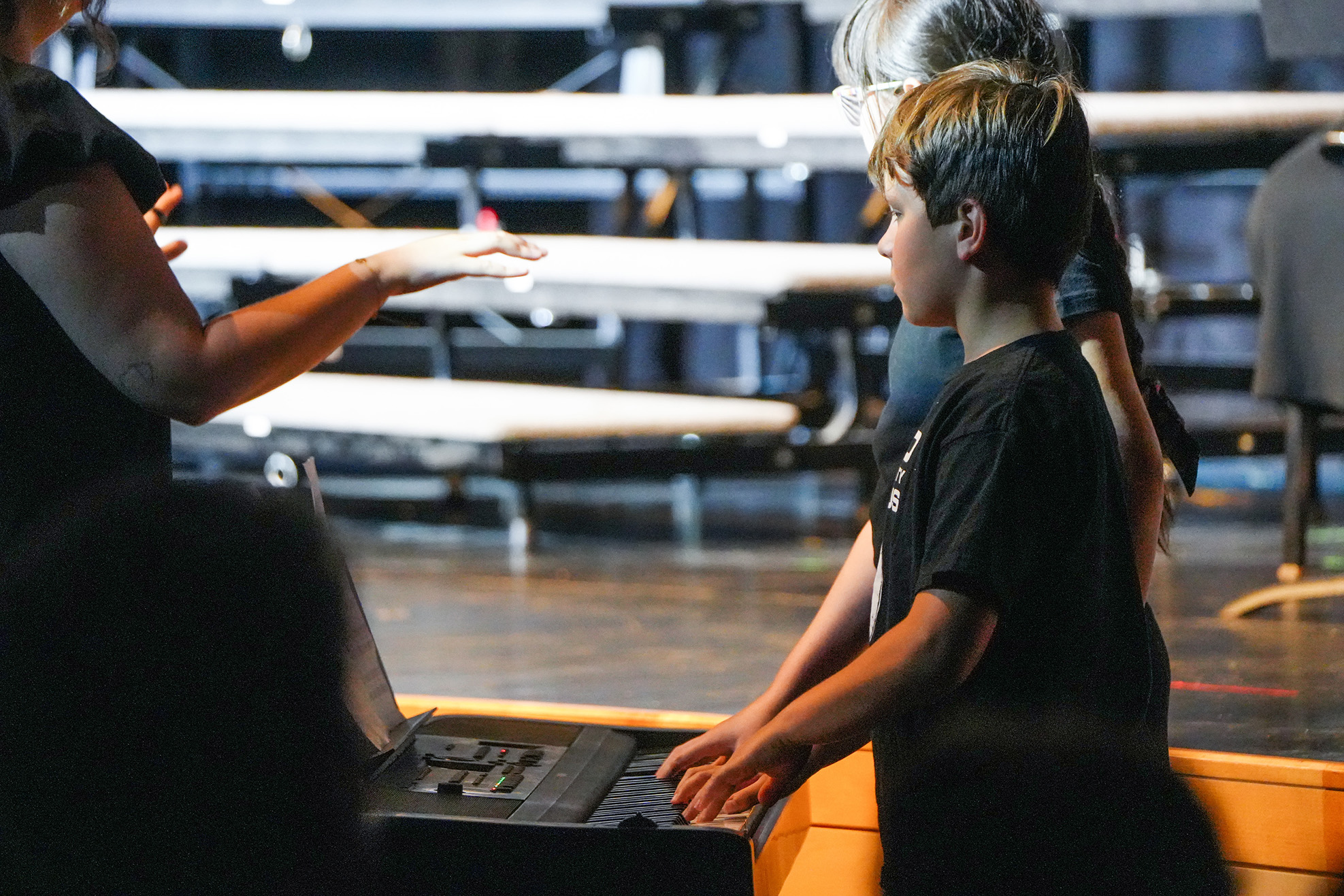 A boy in a black t-shirt plays an electronic keyboard on a stage. A woman in black looks on.