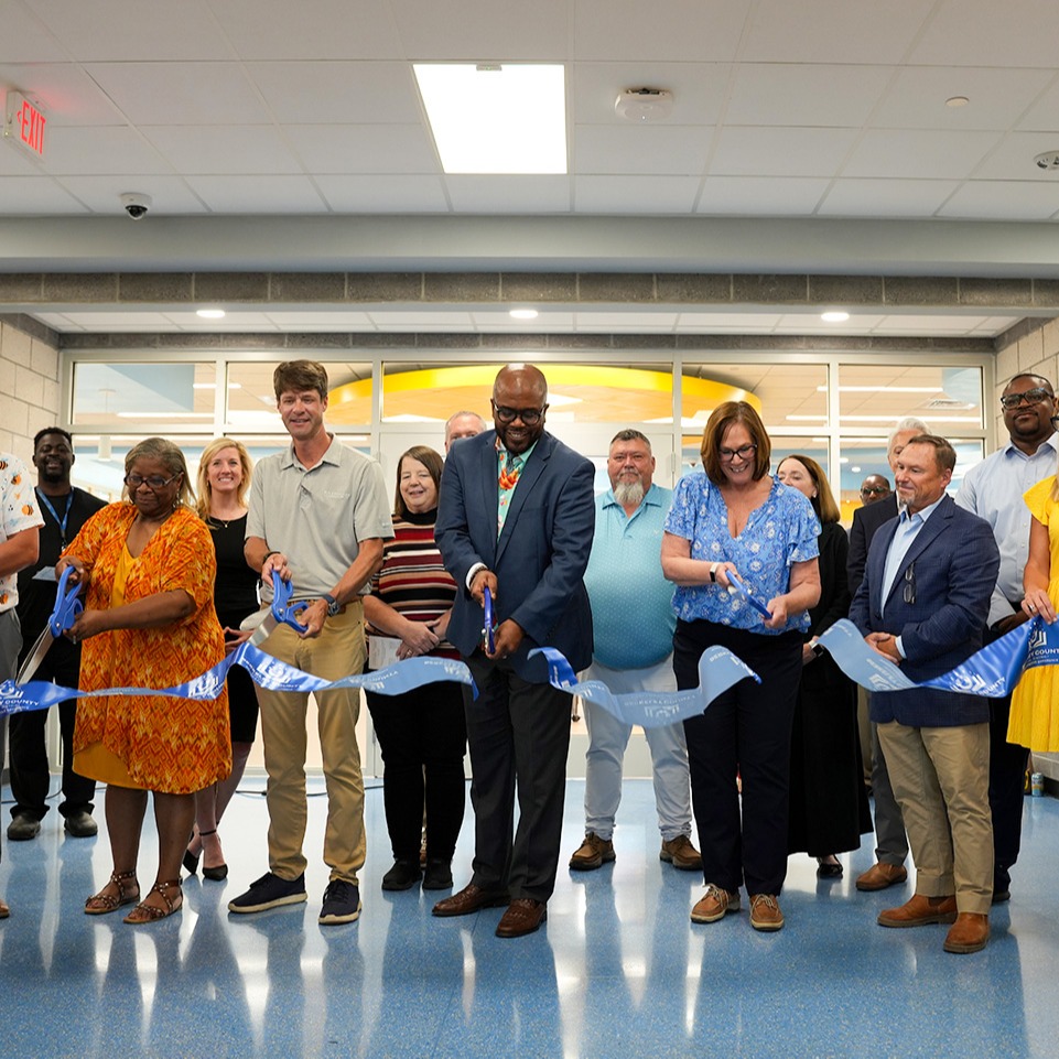 A group of adults cuts a ribbon indoors, surrounded by colorful balloons, with an open doorway in the background.
