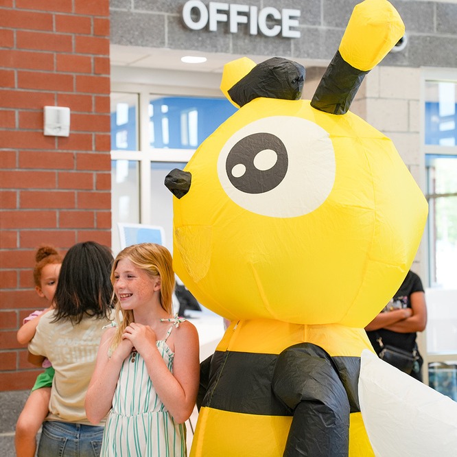 A girl is hugging a bee costume in an indoor area, with people standing behind them.