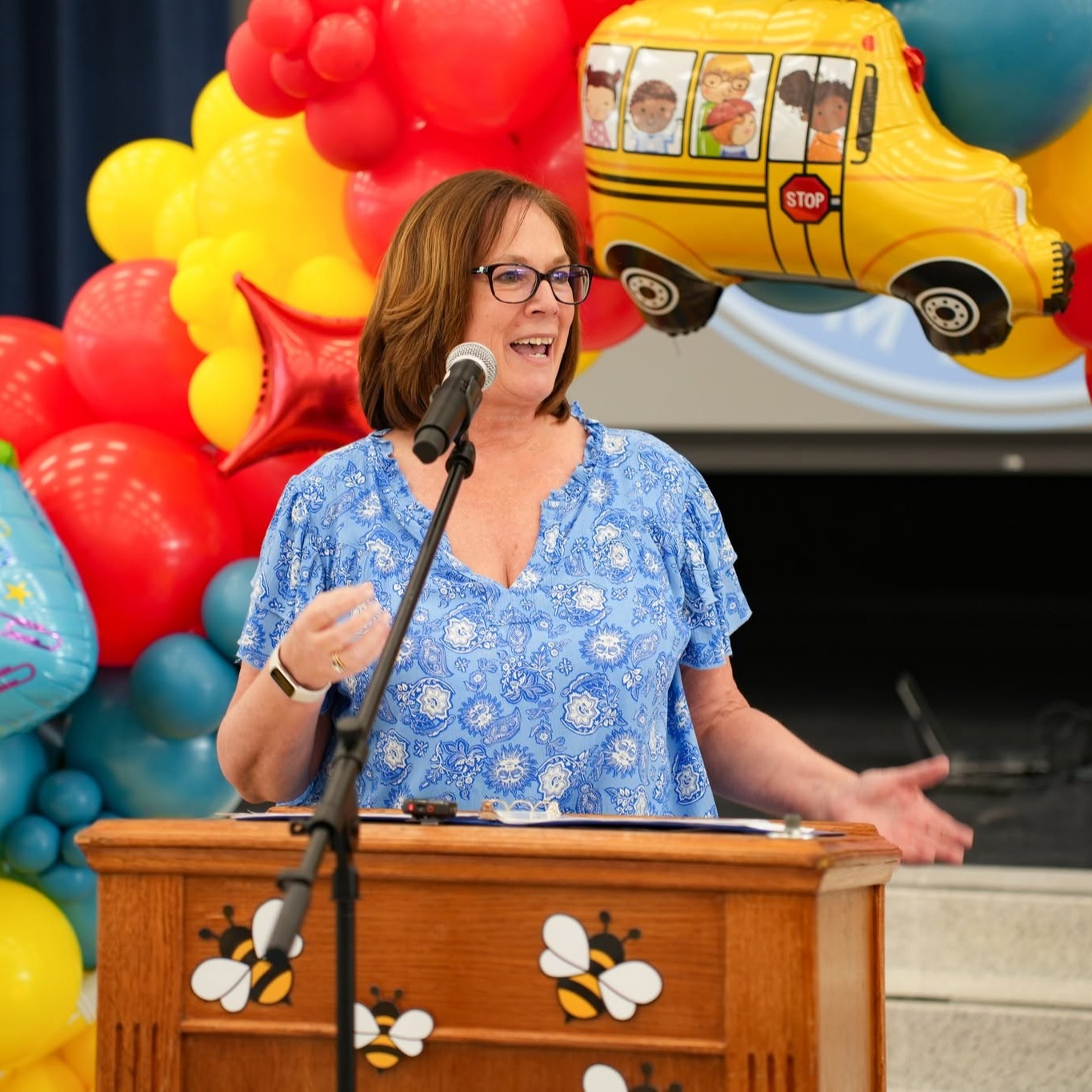 Woman speaking at podium with colorful balloons and a yellow school bus in the background.