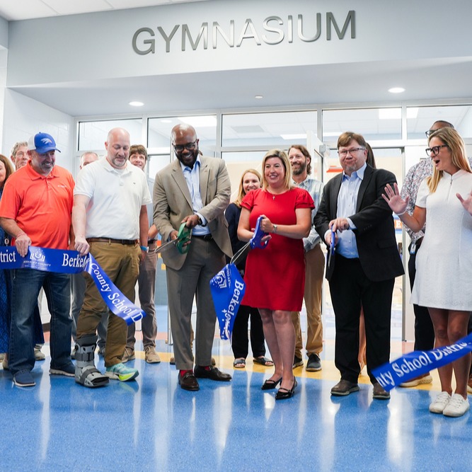 A group of adults stand in front of a ribbon labeled "GYMNASIUM." They appear to be attending a ribbon-cutting ceremony.