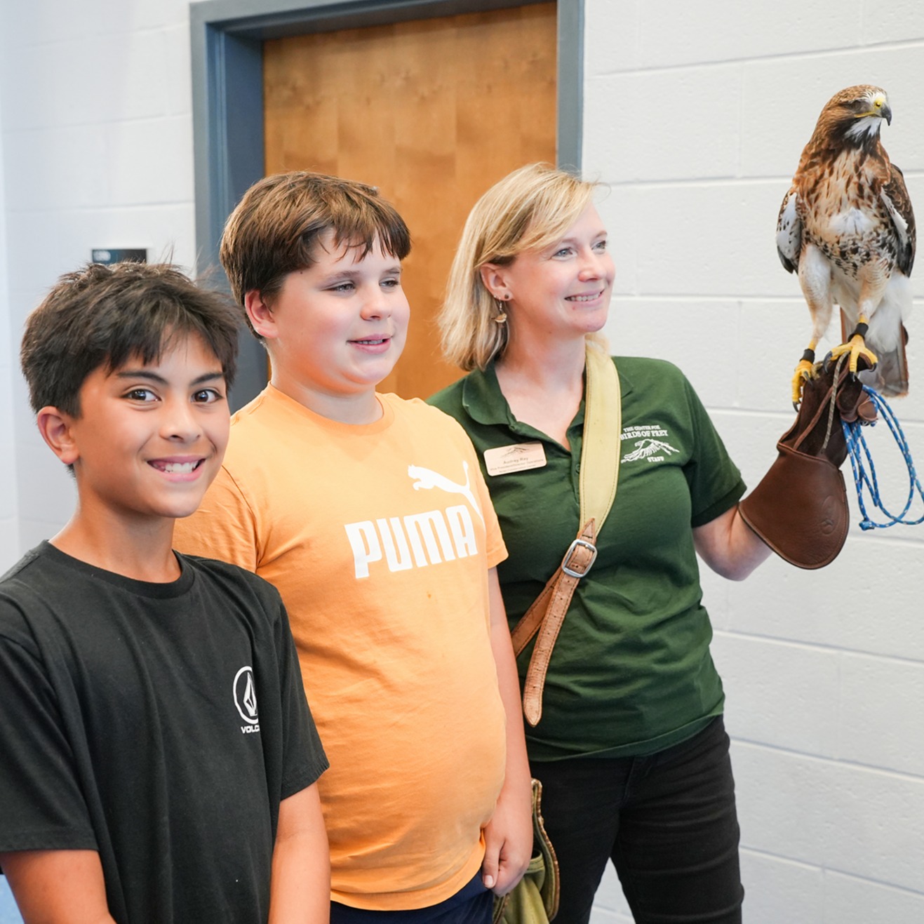 Three children stand next to a woman in a green shirt holding a hawk. Bright balloons decorate the background.