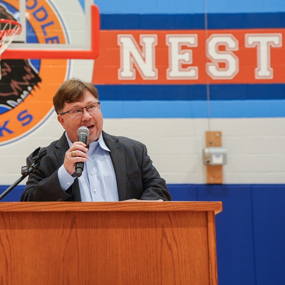 A man speaks into a microphone at a podium. Behind him, a wall displays the word "NEST" and a Hawks logo.