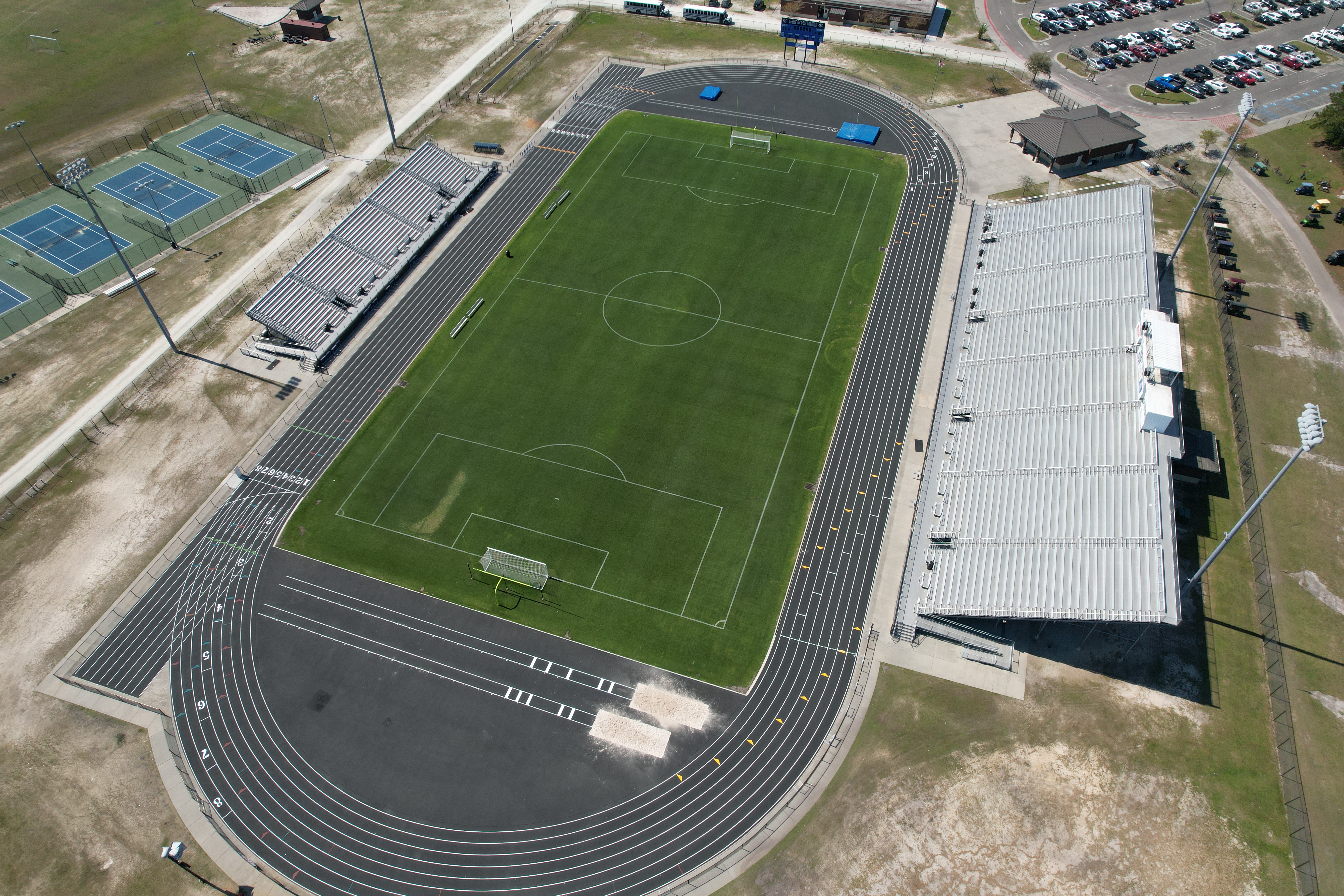 A stadium with a green field, running track, and large seating area. A parking lot with cars is in the background.