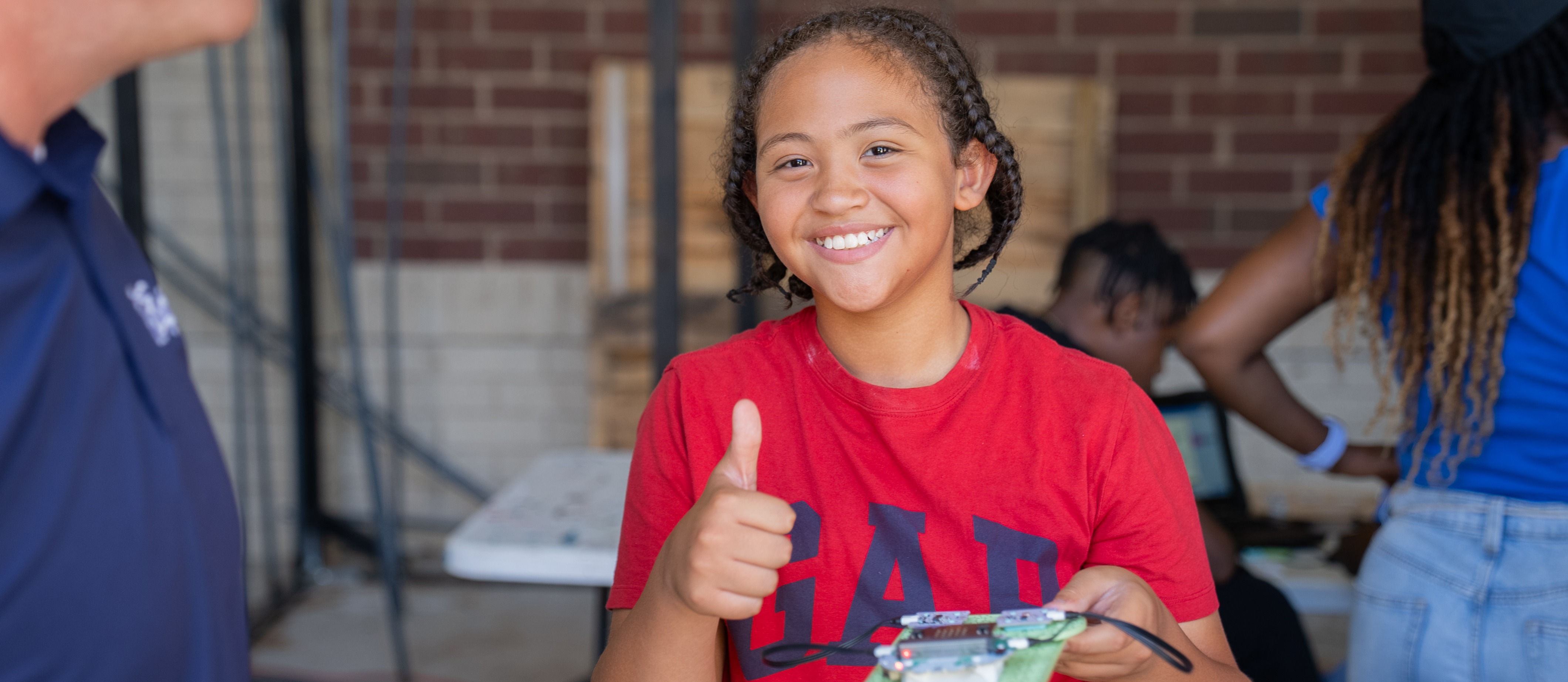 A young girl in a red shirt gives a thumbs up, standing in front of a brick wall.