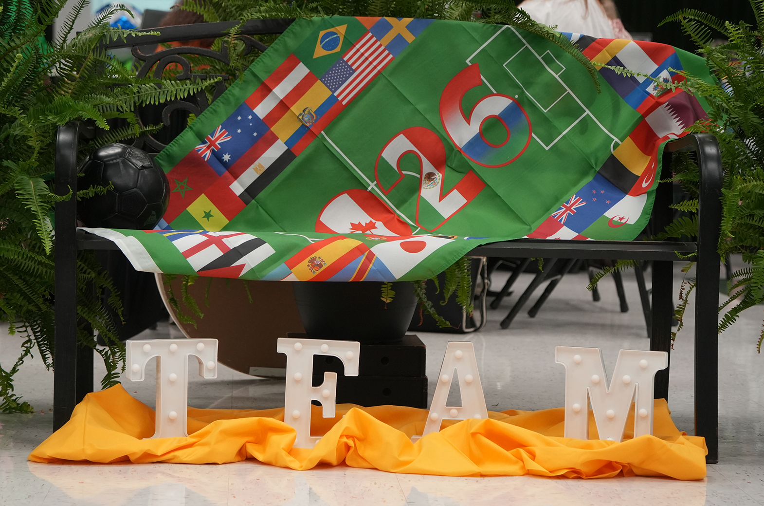 Displayed is a bench with a blanket featuring flags from various countries and a soccer field, accompanied by lighted letters spelling "TEAM" on a yellow cloth.