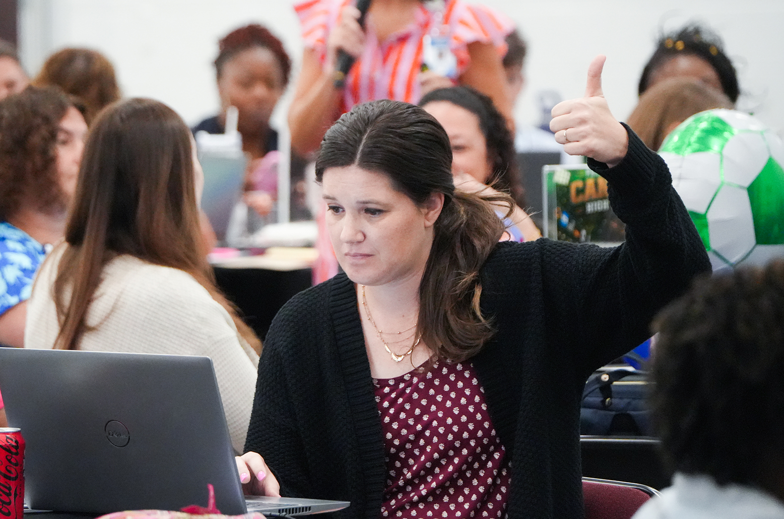 Woman seated at a laptop, giving a thumbs up, surrounded by other people sitting at desks.
