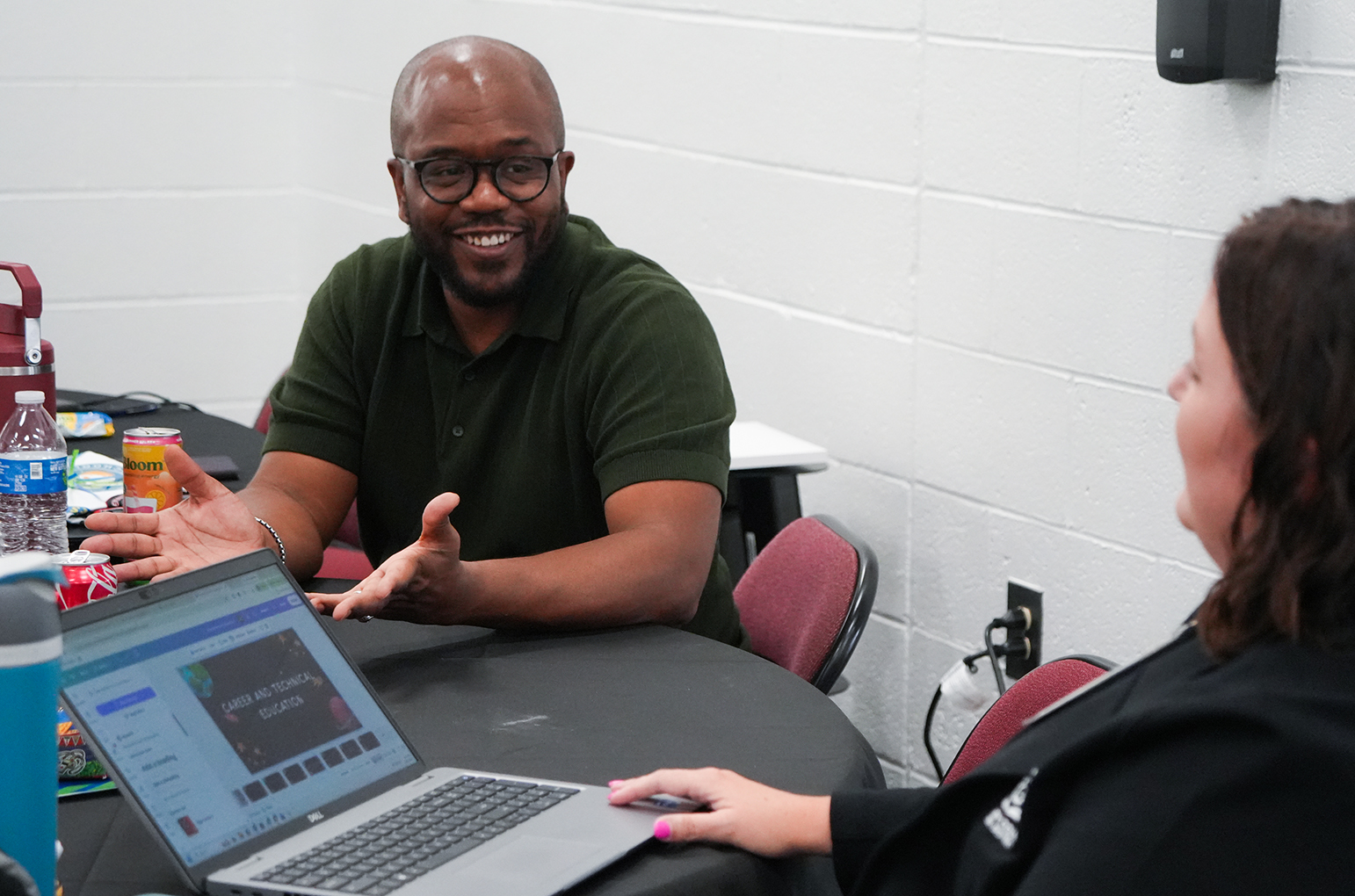 Man in green shirt sitting with woman at table, smiling and gesturing while looking at open laptop.