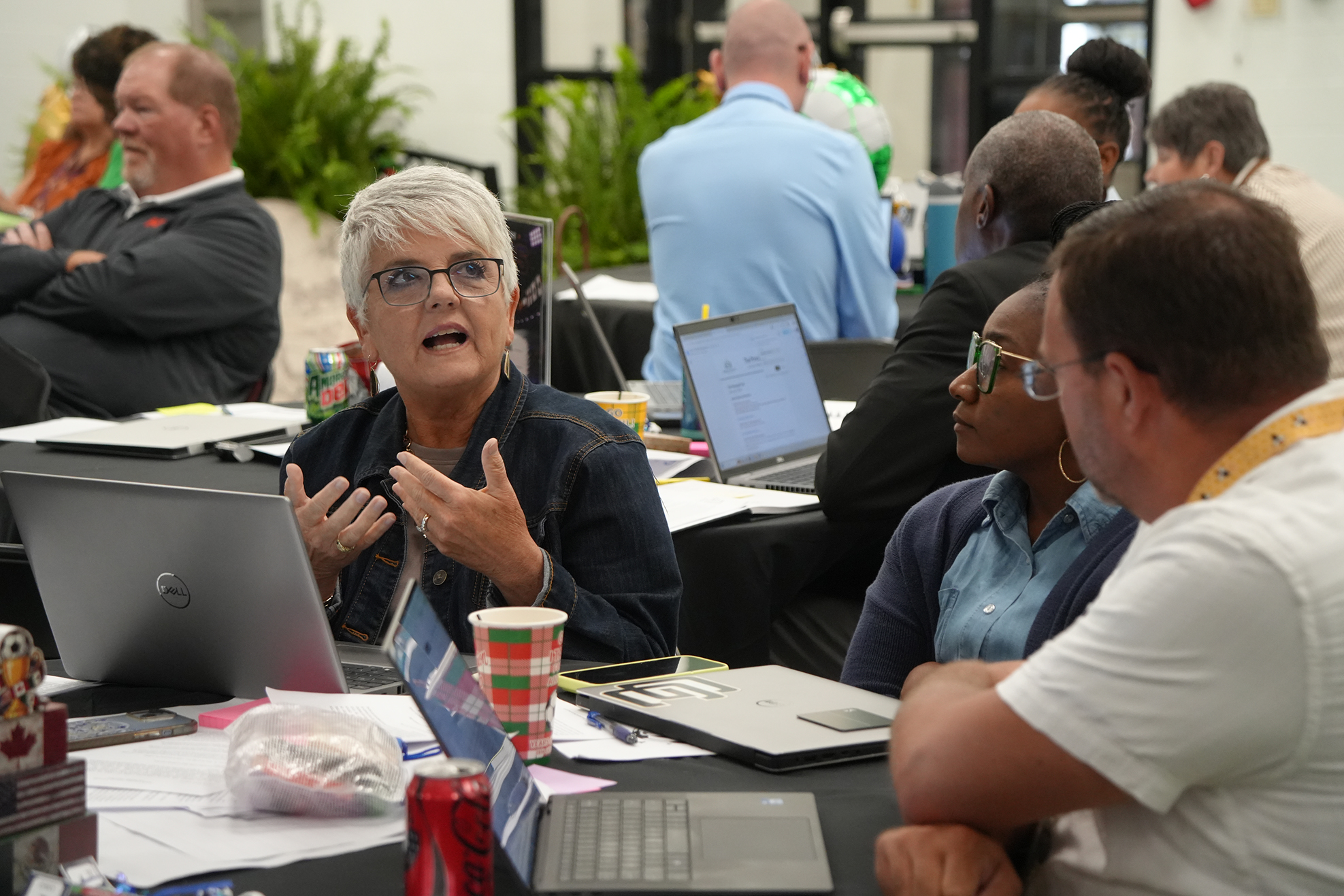 A group of people sit at tables with laptops. A woman speaks while others listen.