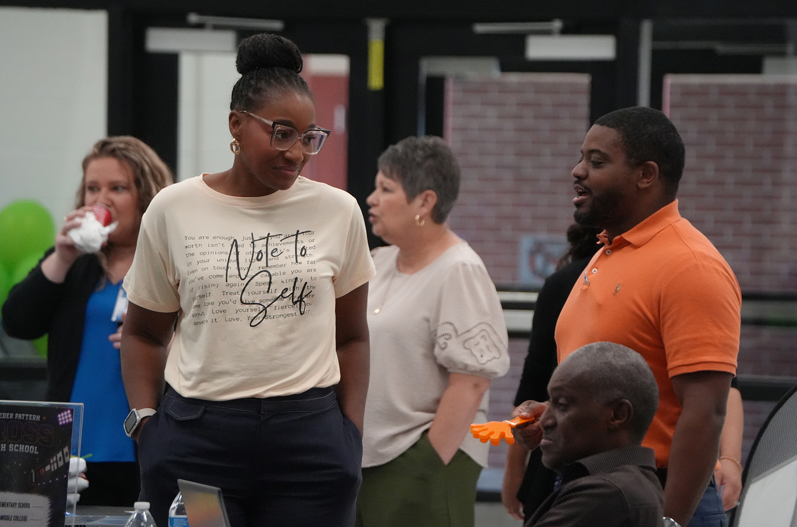 A group of people stand in a room; a woman with glasses wears a white shirt. A man in orange talks to a seated man.