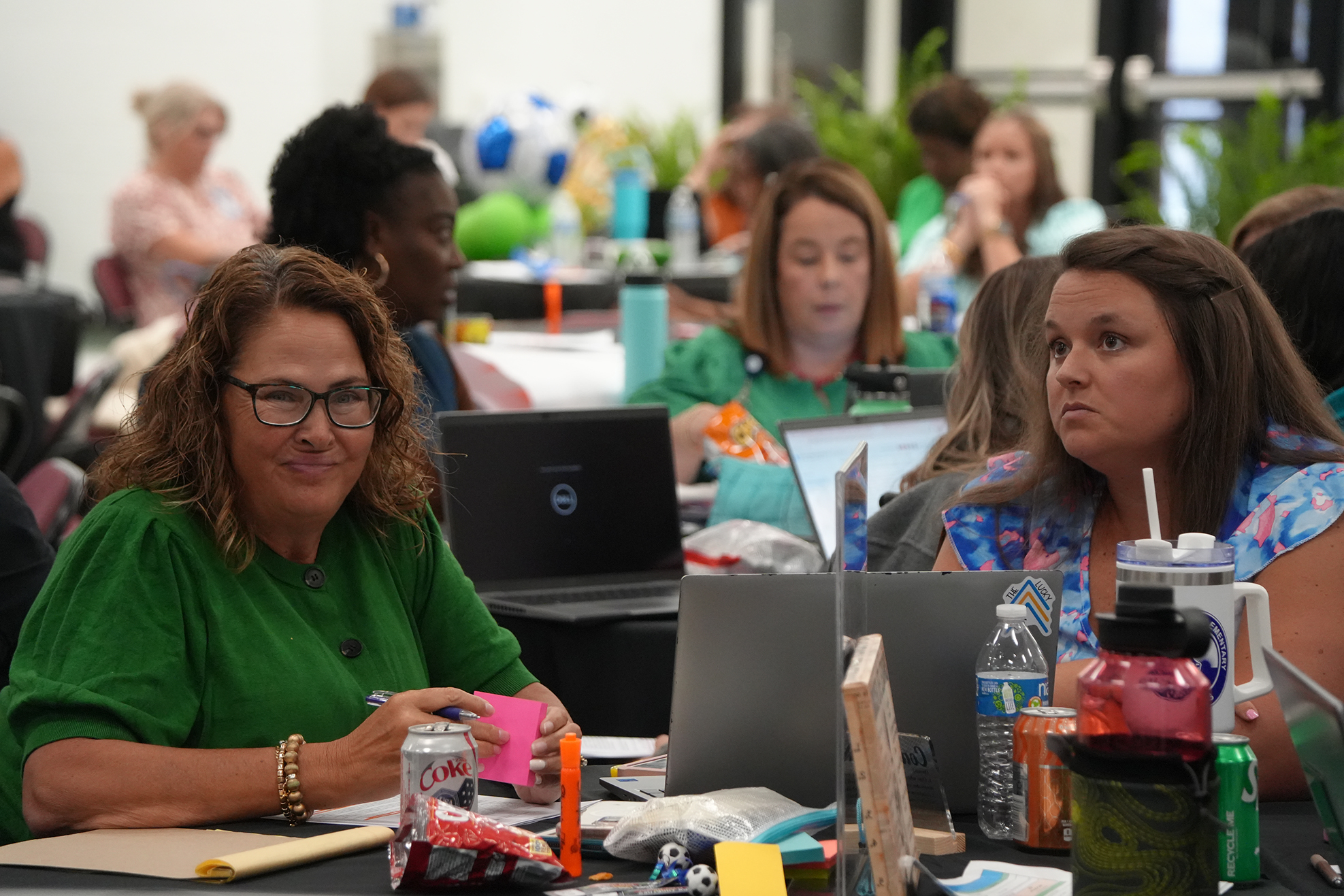 People sit around tables with laptops and water bottles; one woman wears a green shirt, another a blue shirt.