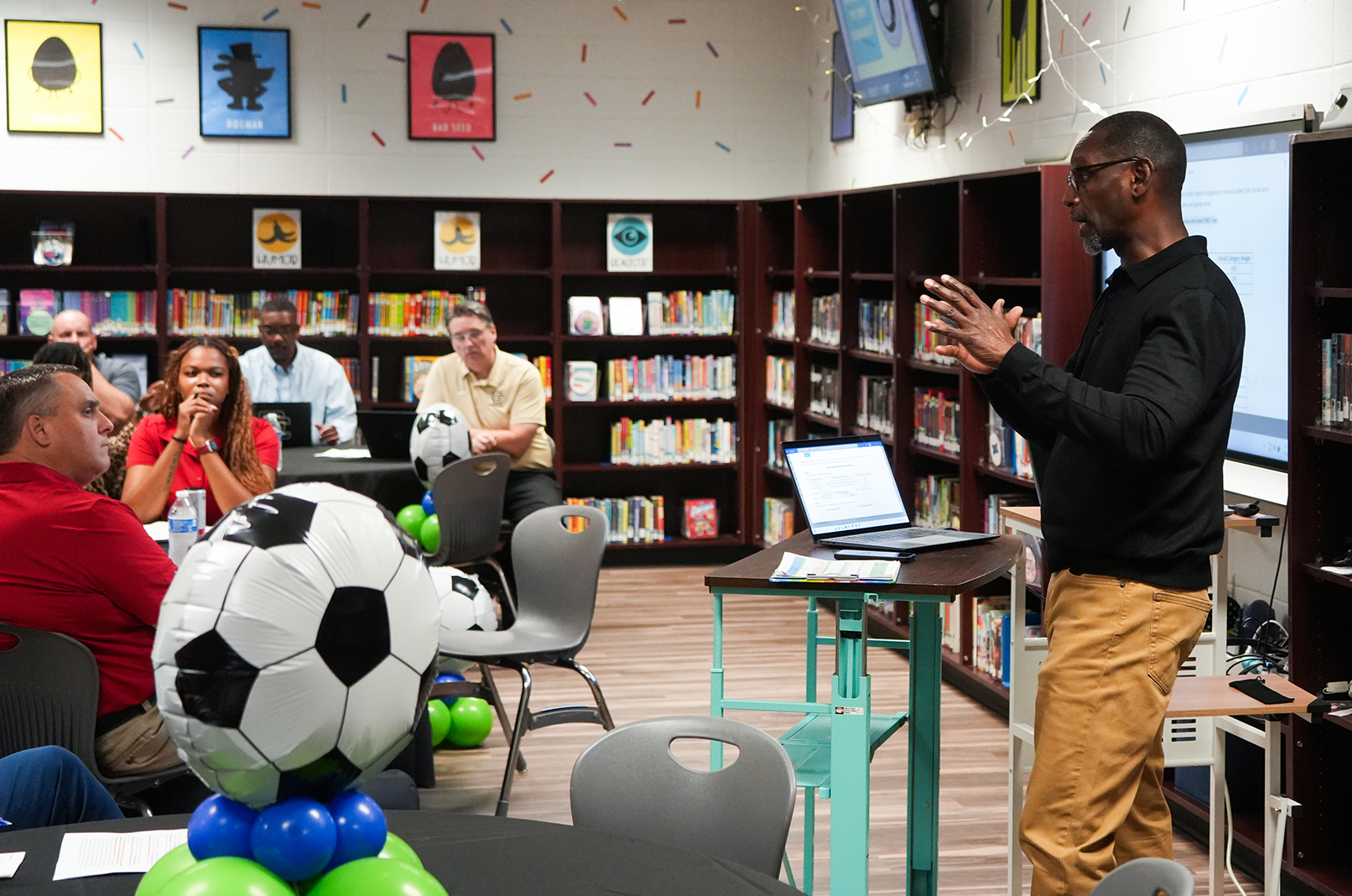 A man speaks to a seated audience in a library. Shelves with books are in the background. A soccer ball decoration is on a table.