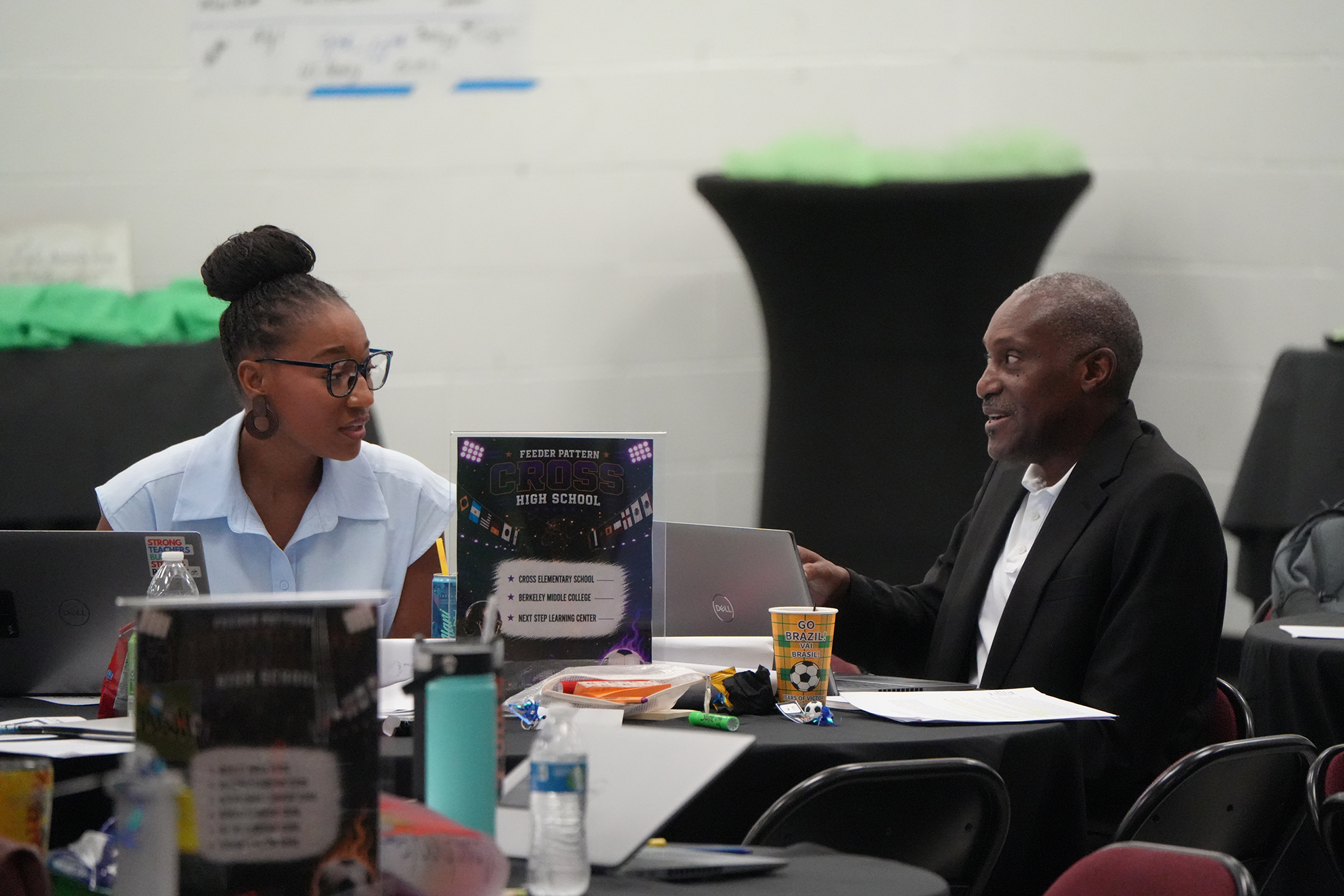 A woman and man seated at a table with laptops, a book, and a cup.