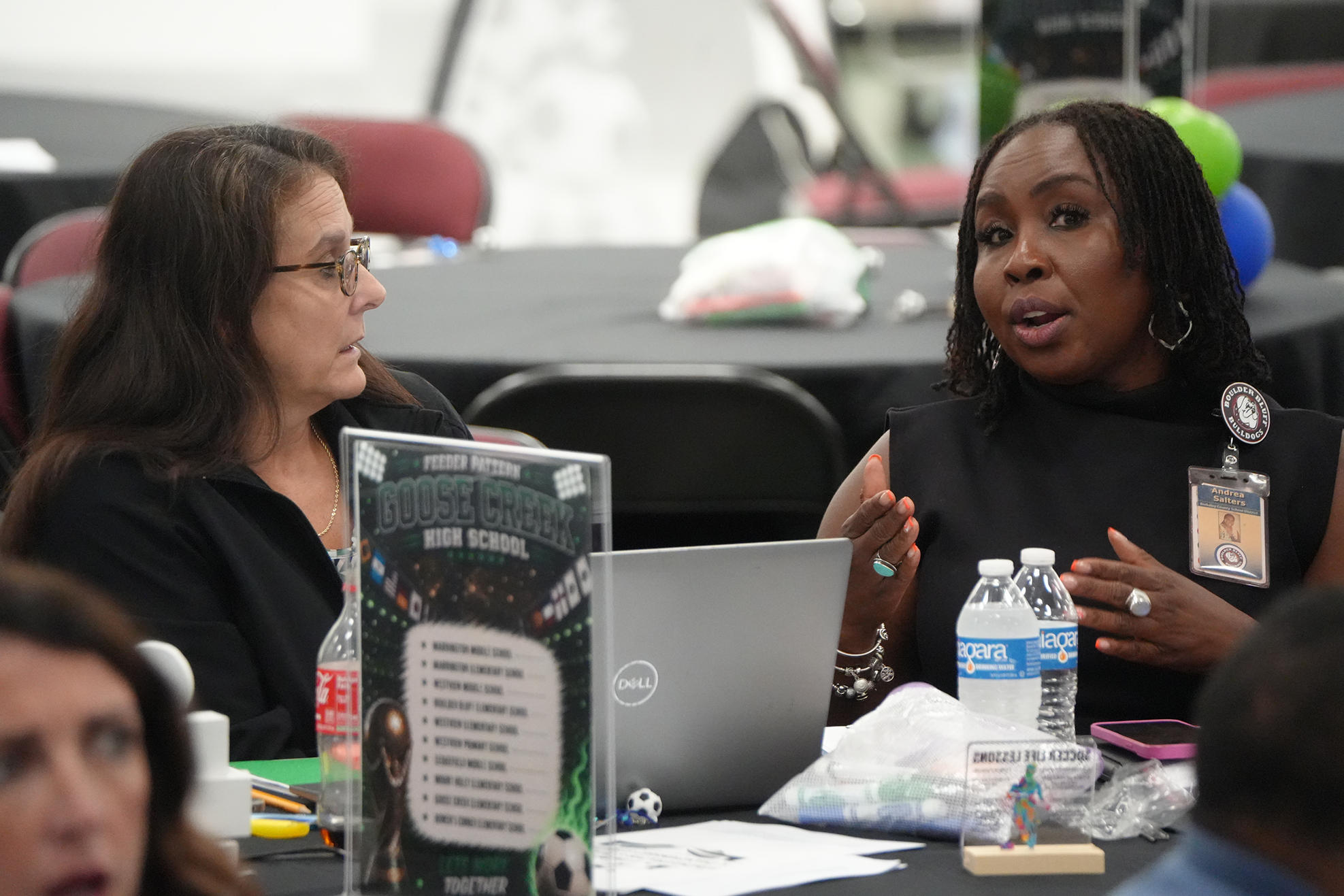 Two women at a table with a laptop, one speaking and the other listening, surrounded by papers and water bottles.