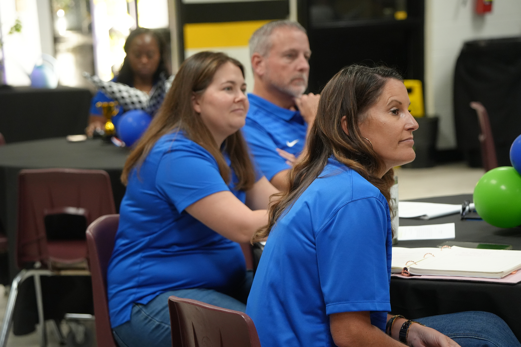 Four people in blue shirts sit around a table with balloons; two women face forward and two men sit behind.