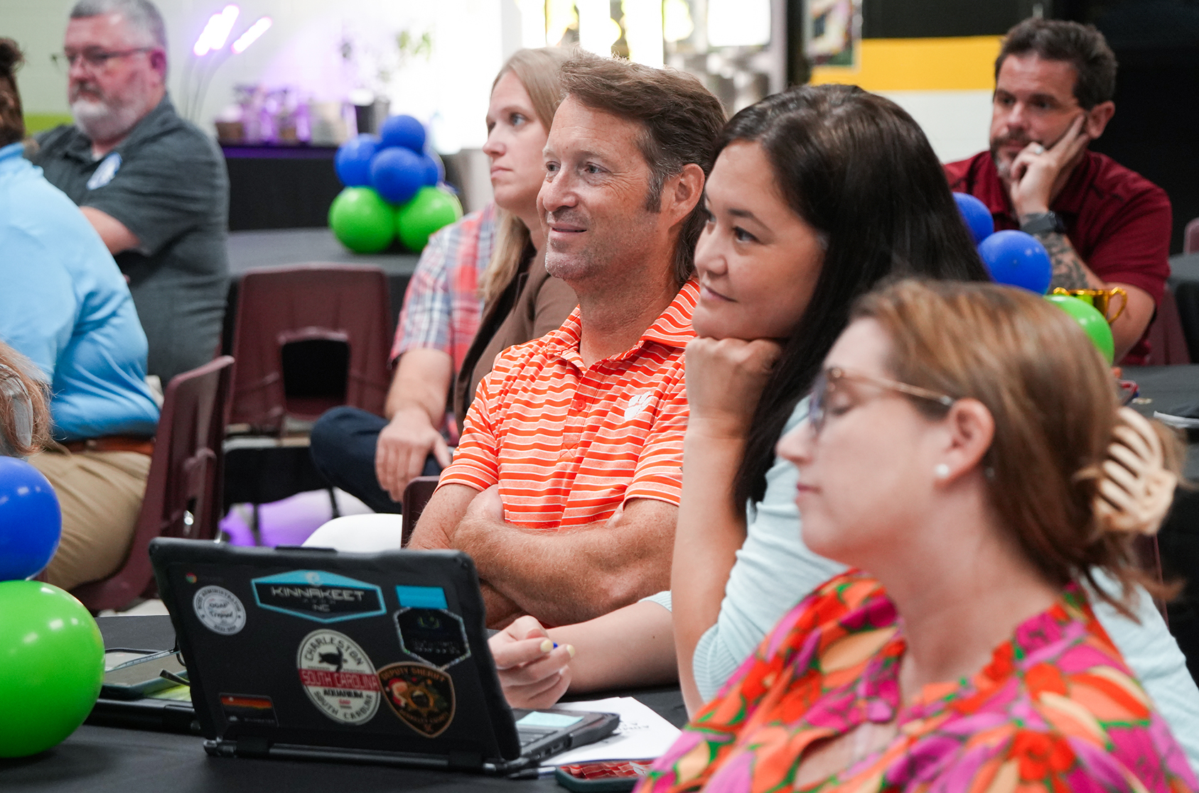 People sit in a classroom with balloons, one with a laptop displaying stickers, two looking at it.