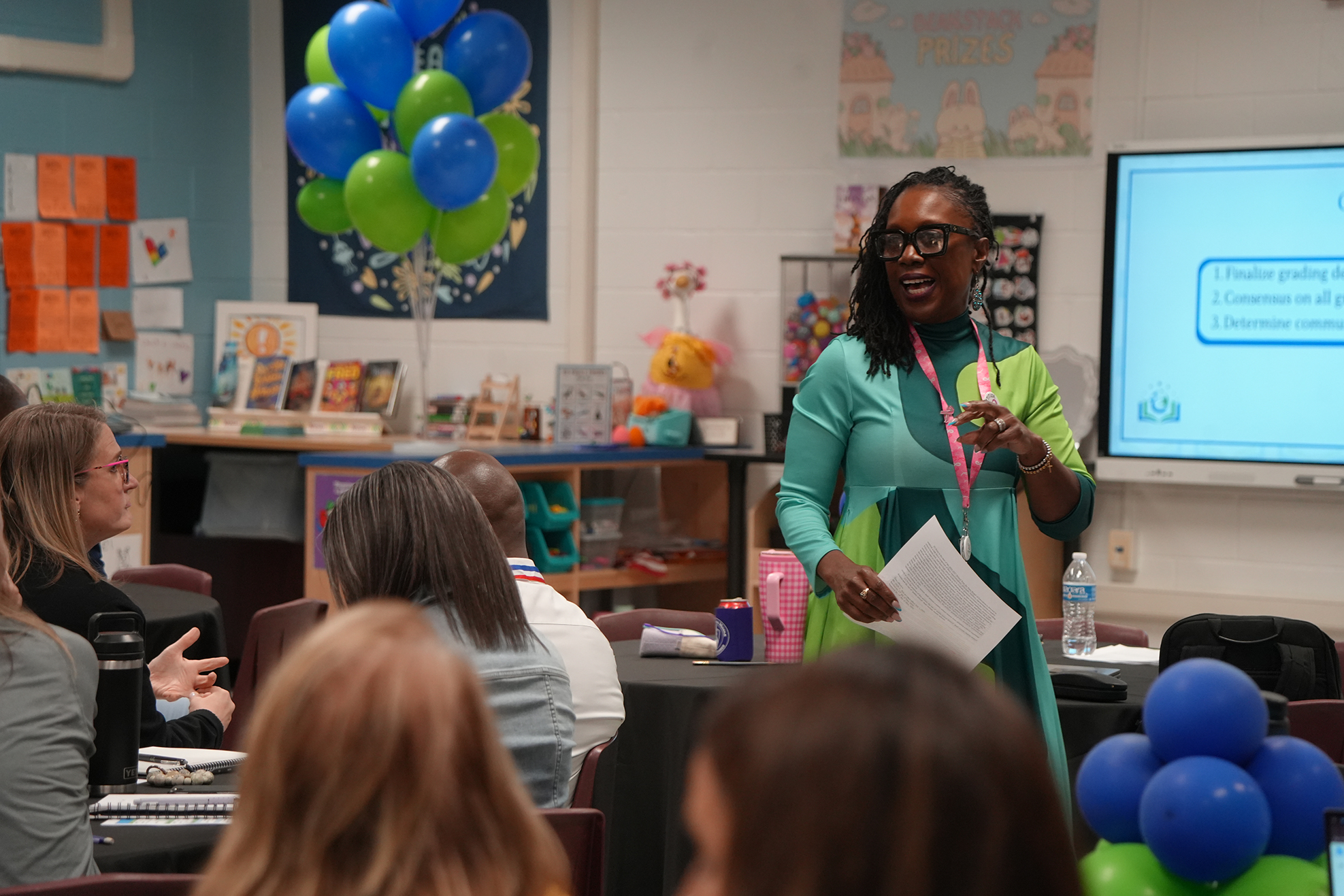 A woman speaks to an audience in a classroom with a large monitor, balloons, and a table.