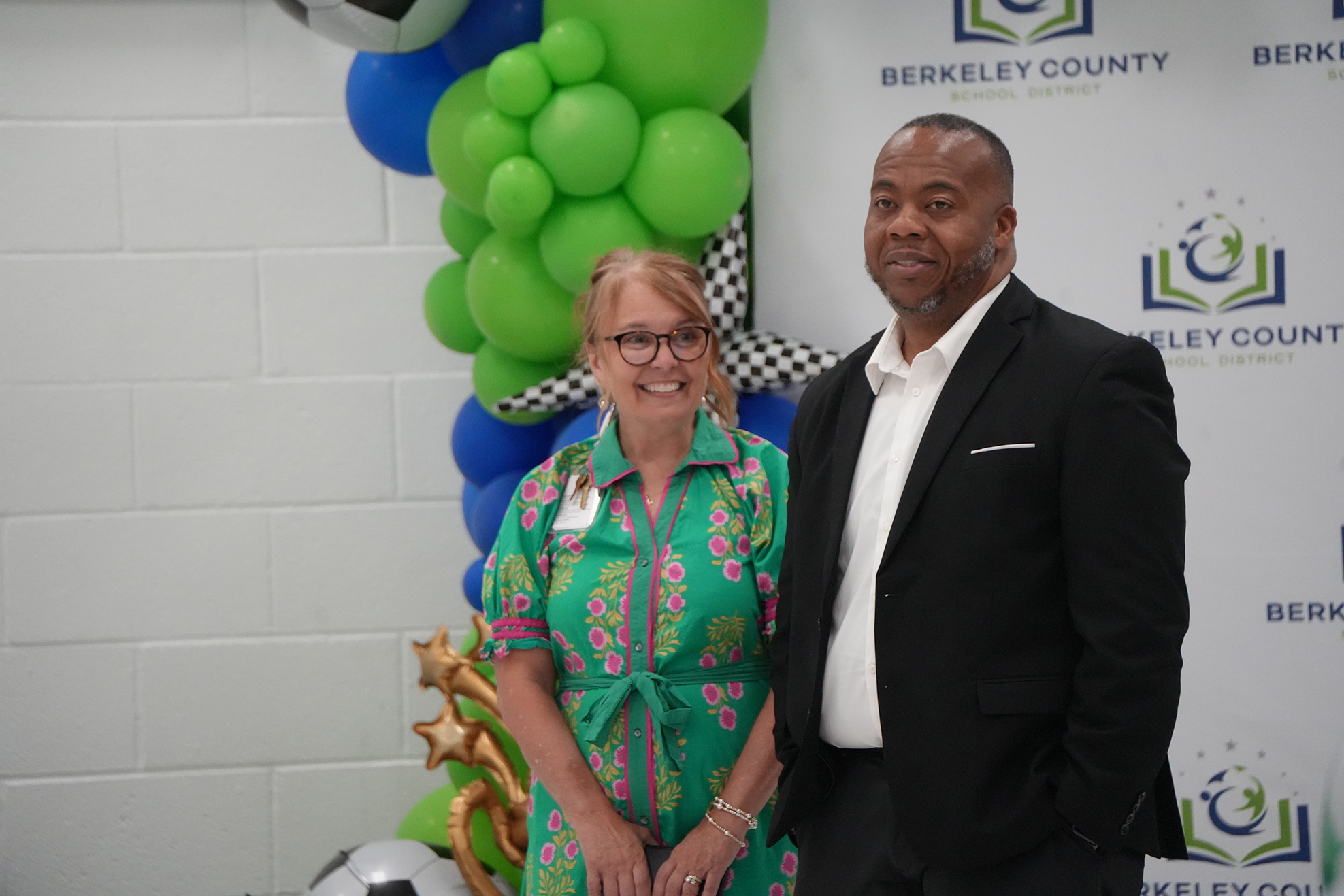 A man and a woman, both dressed formally, standing in front of a wall with balloons and a logo.
