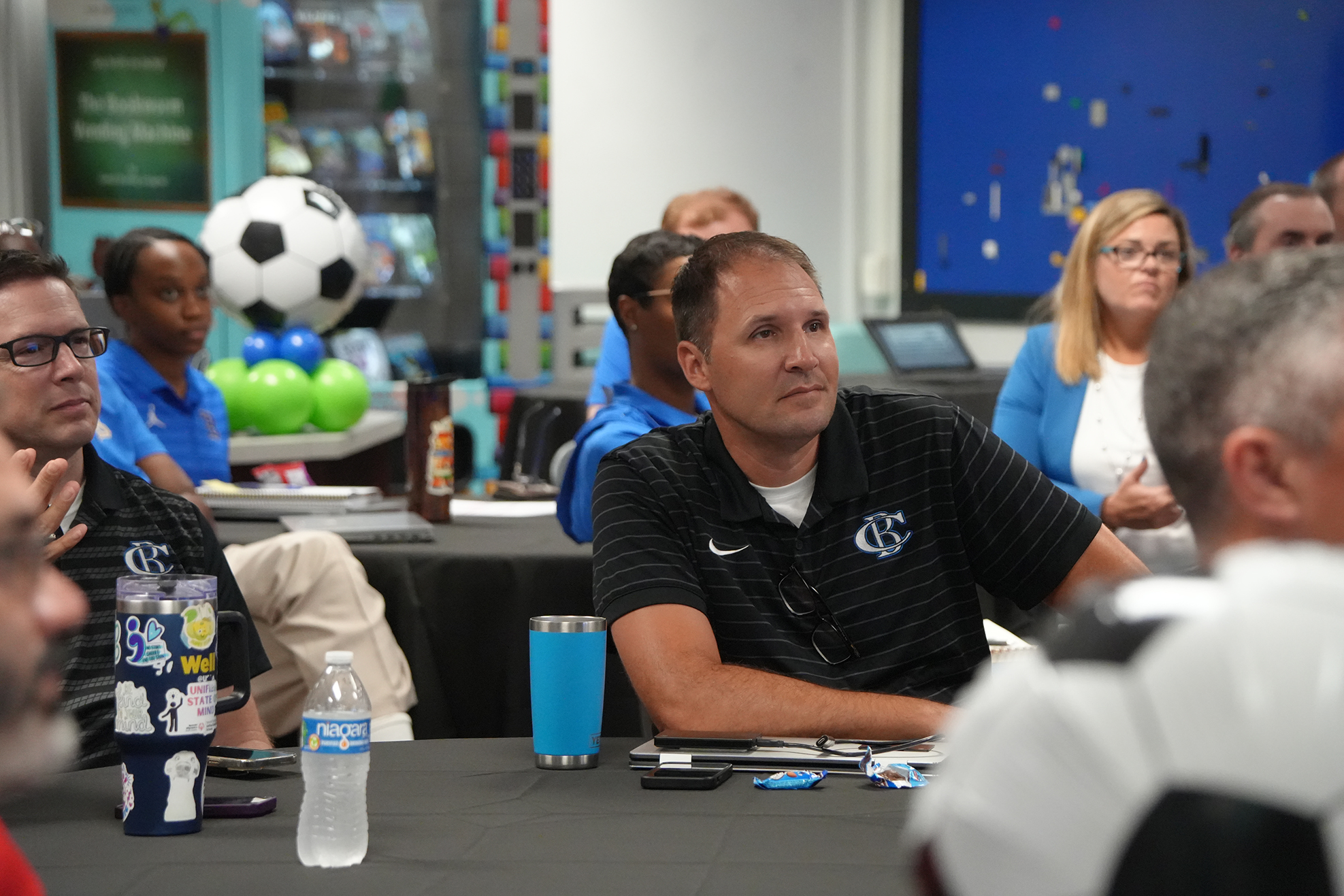 People in a room with tables, one wearing a black shirt with text and logo. A soccer ball hangs in the background.