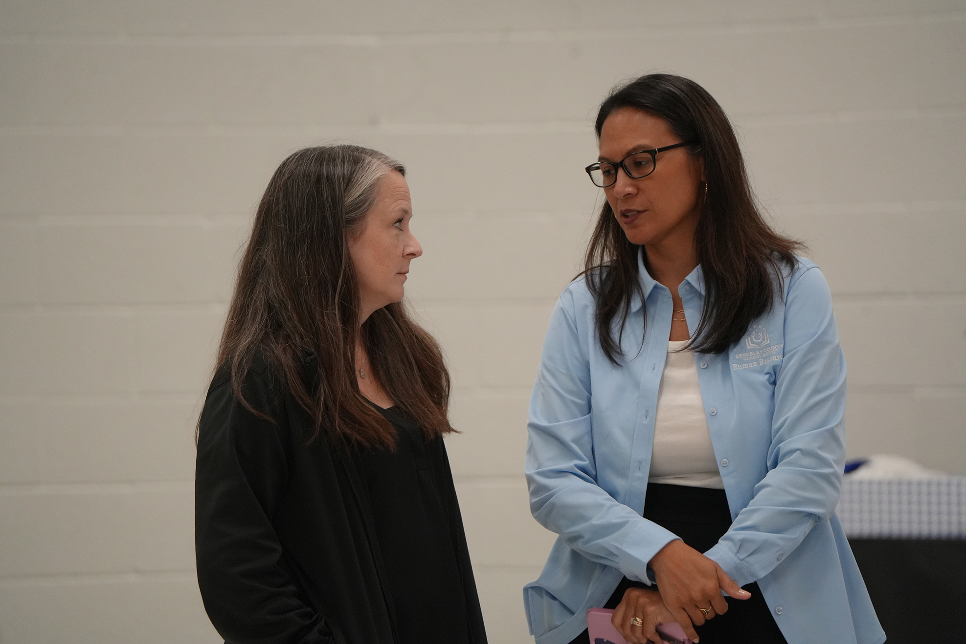 Two women face each other in a room with a white background. One wears a blue shirt and glasses, the other a black top.