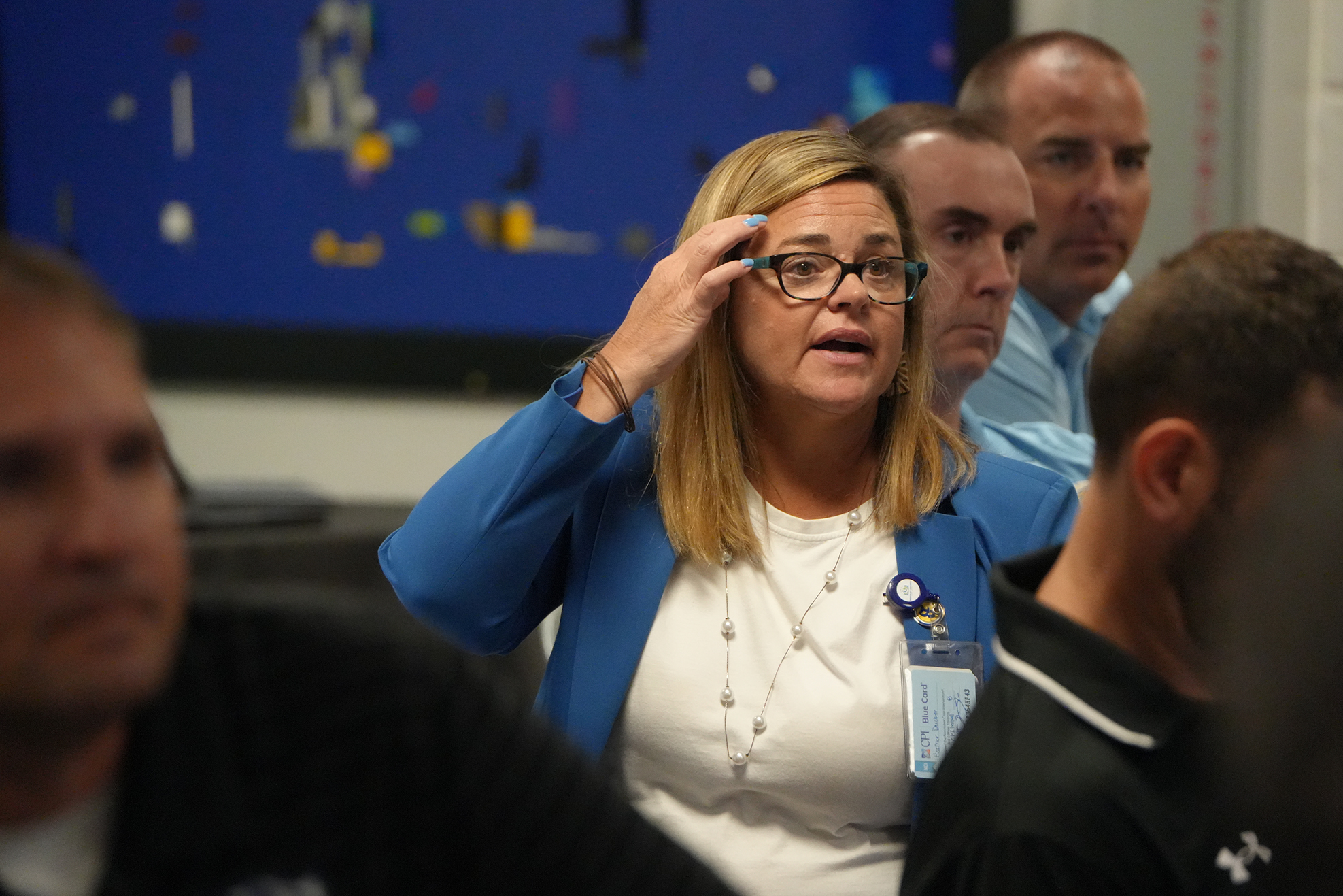 A woman with blonde hair wearing glasses and a blue blazer speaks in a meeting with others.
