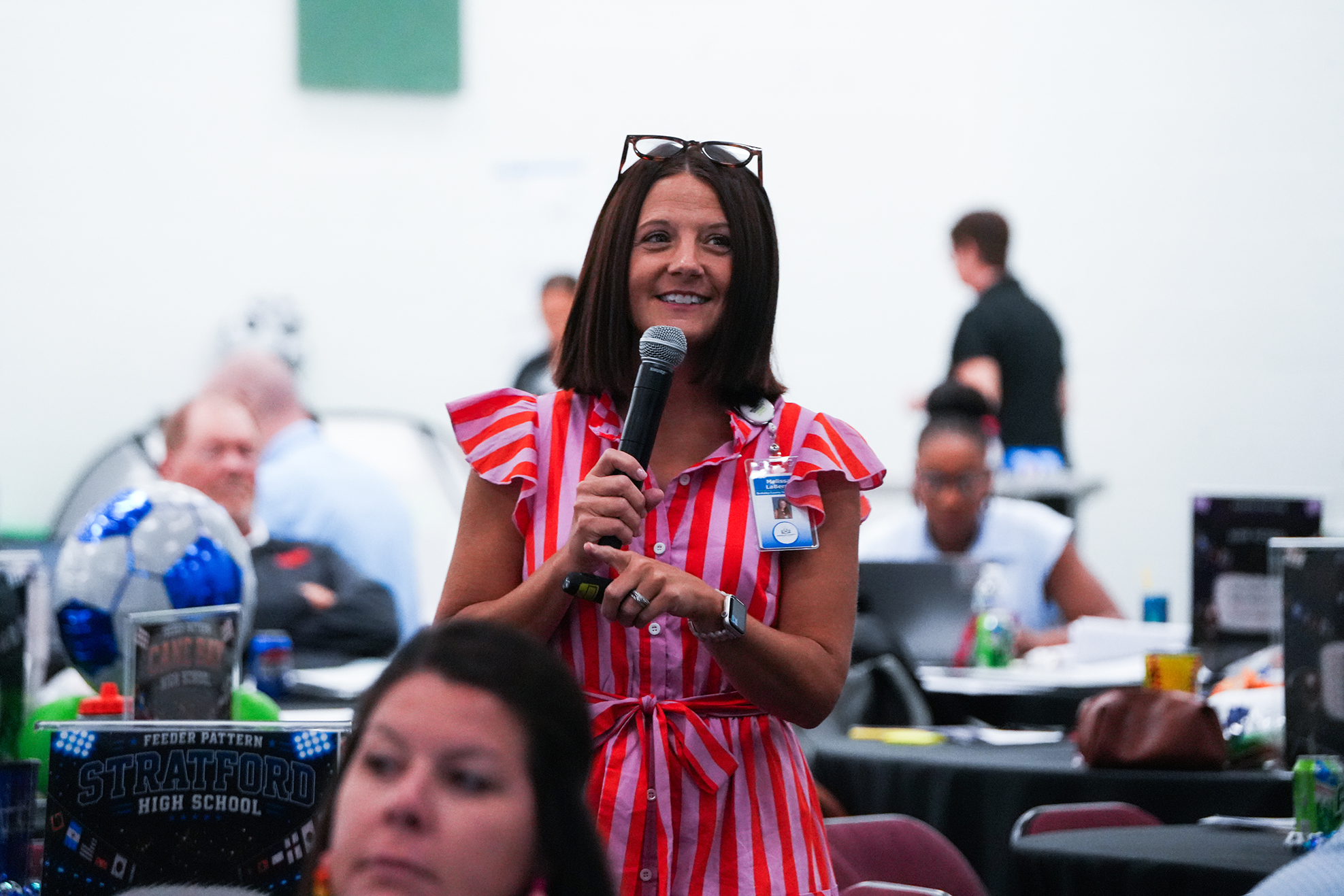 Woman in a red dress speaking into a microphone at an event; audience seated at tables behind her.