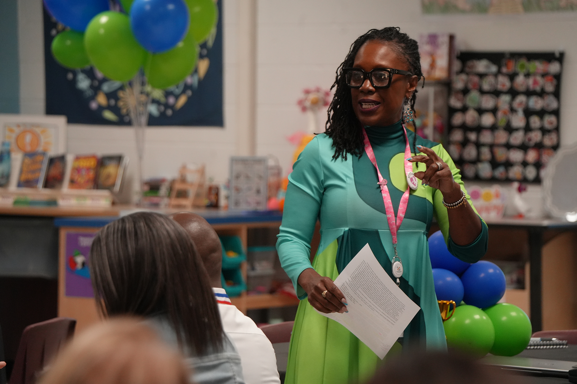 A woman in a green dress holds a paper while addressing a classroom audience. Behind her, colorful balloons and a bulletin board are visible.