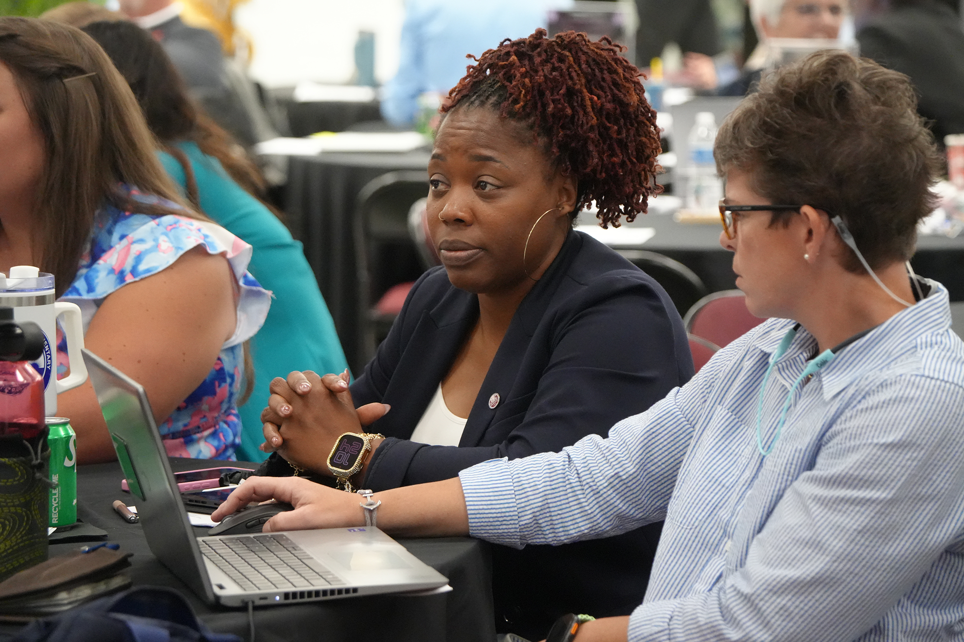 Two people sitting at a table in a conference room, one using a laptop, the other wearing glasses.