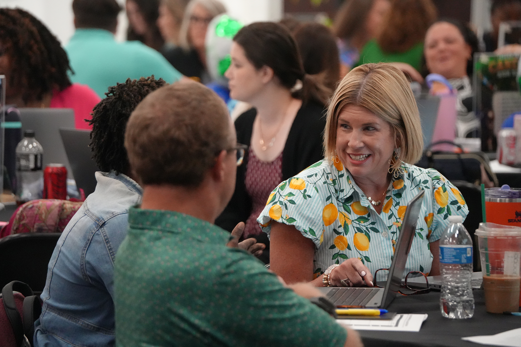 Group of people in a room with laptops. A woman with blonde hair is smiling and looking at a man.