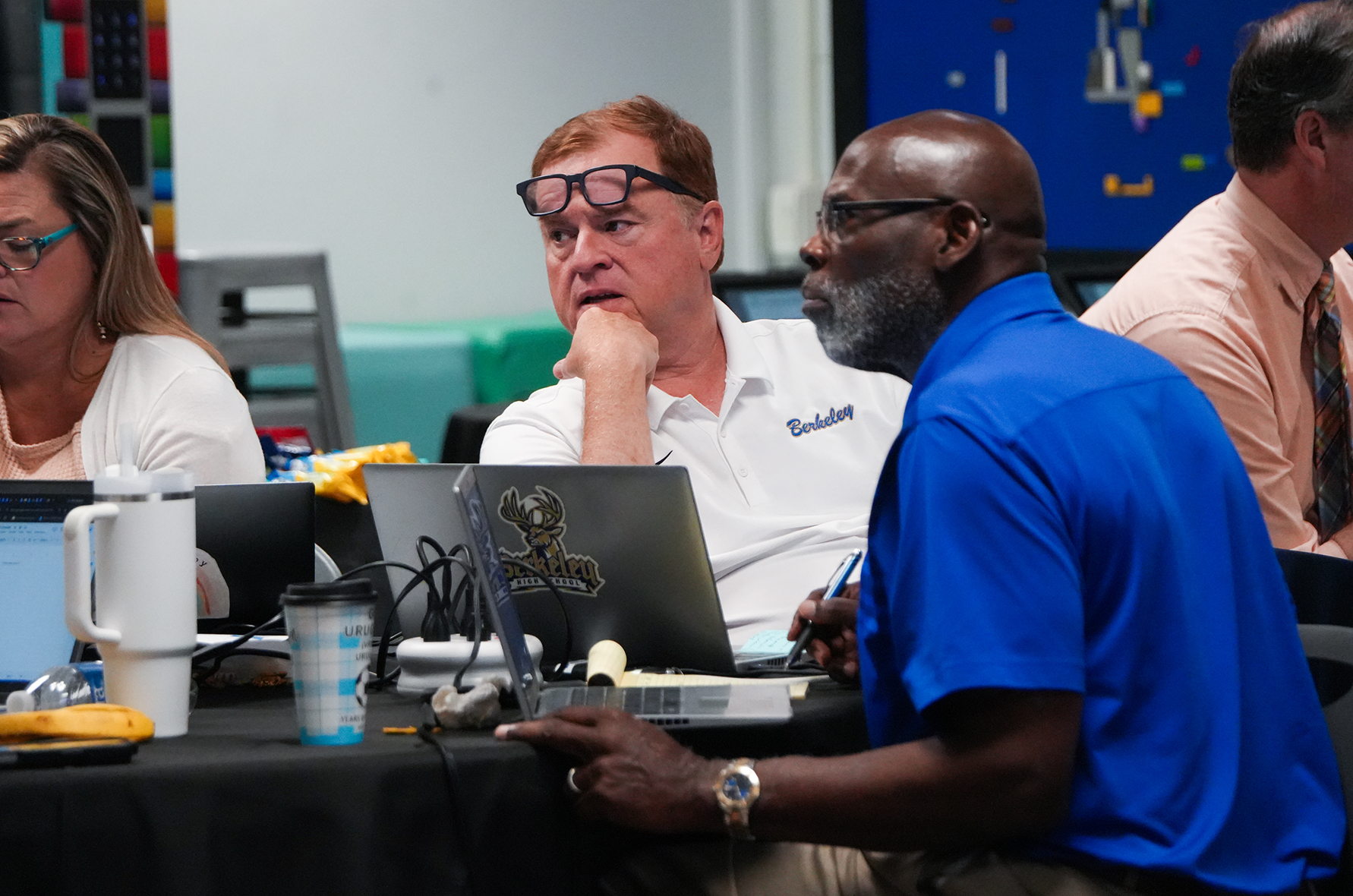 A group of people seated around a table, each engaged with laptops. One man in blue wears a watch, and others wear glasses.