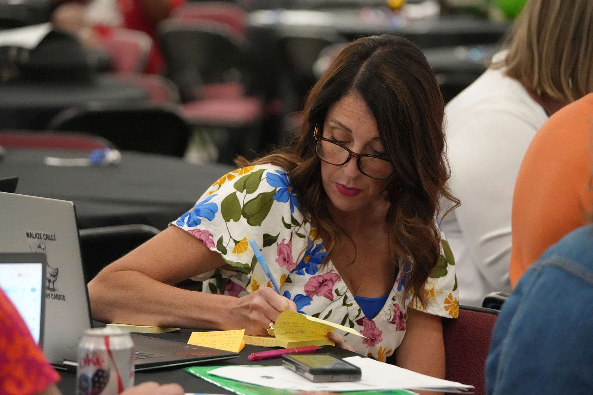 Woman wearing glasses sits at a table, writing on a sticky note with a pen.