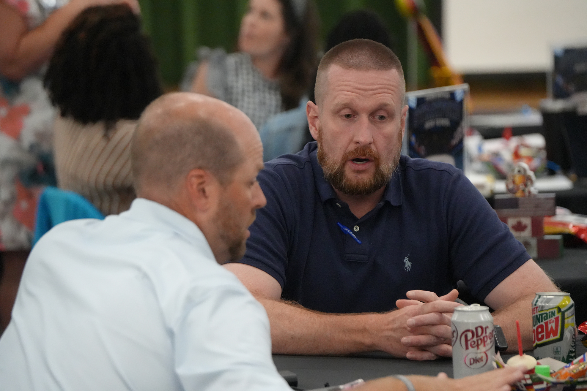 Two men talk in a room with people in the background. One man holds a can of Pepsi.