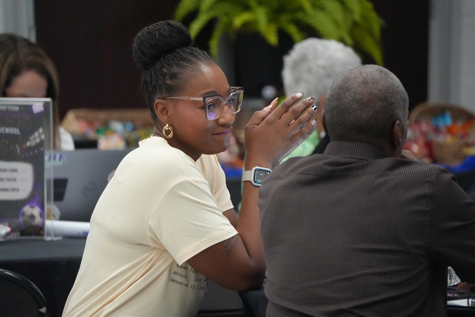 A woman with glasses speaks to a man with a dark jacket at a table with computers.