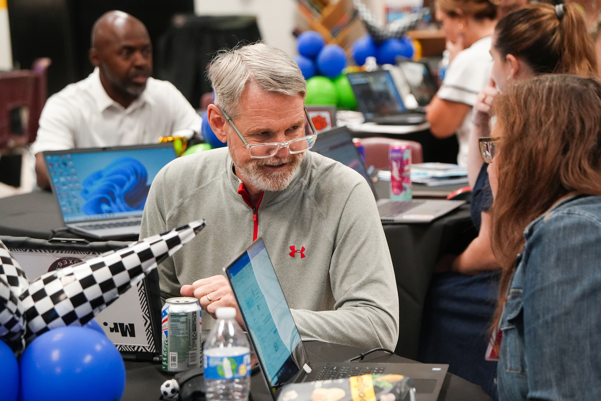 Several people sit around a table with laptops; a man in glasses looks at his screen while a woman speaks nearby.
